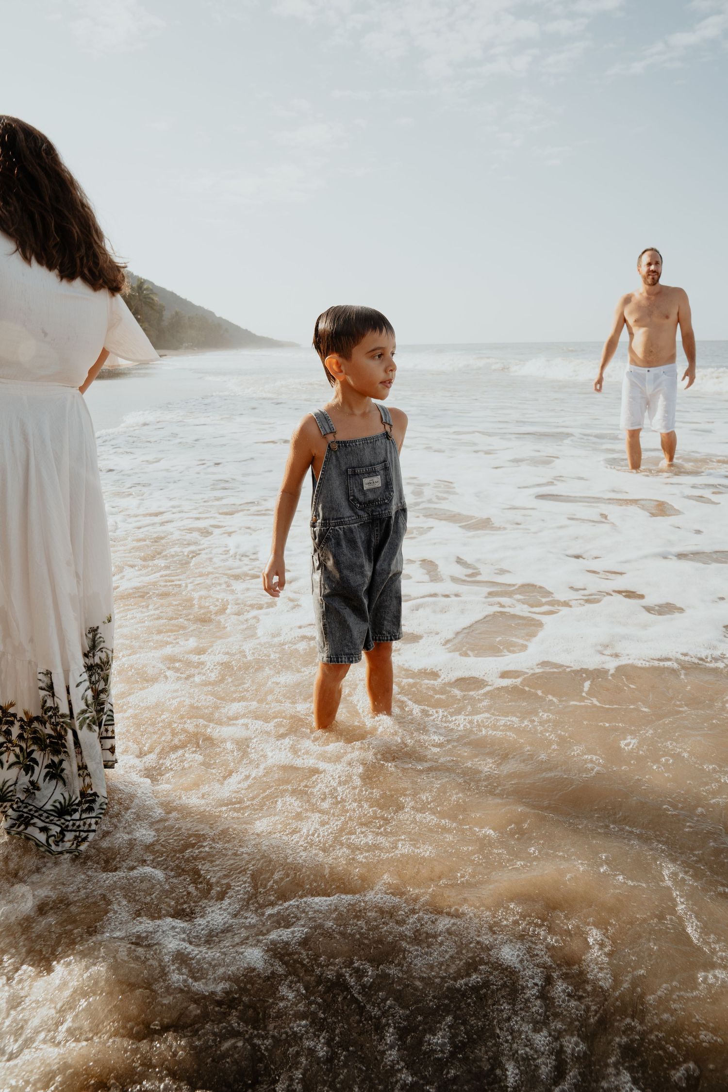 Young child in denim overalls standing in ocean waves on a sunny beach with parents nearby.