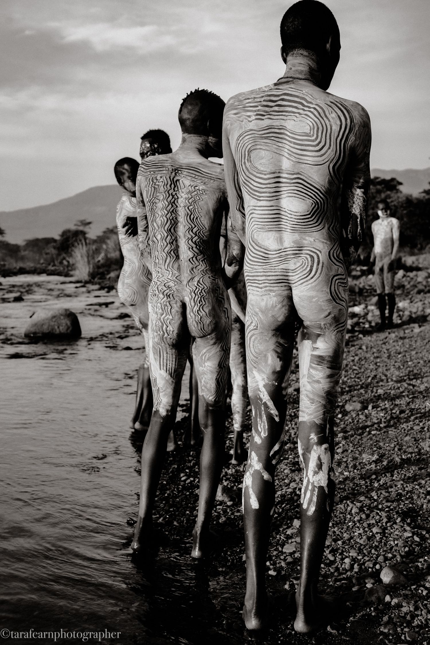 Black and white photo of tribal men with intricate body paint walking toward a rocky river.