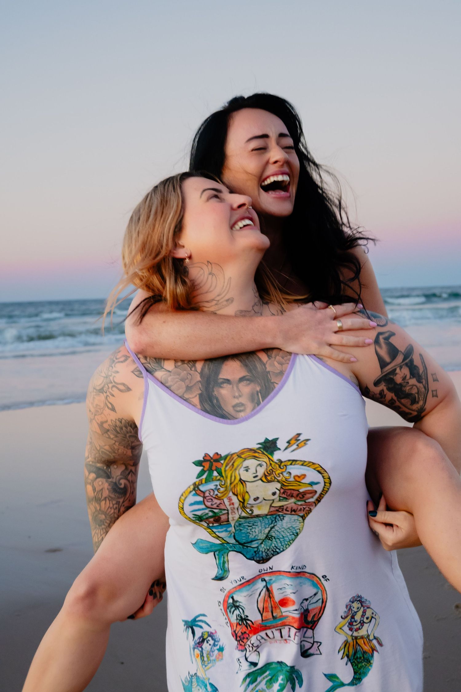 Two laughing tattooed women sharing a piggyback ride on the beach at sunset.