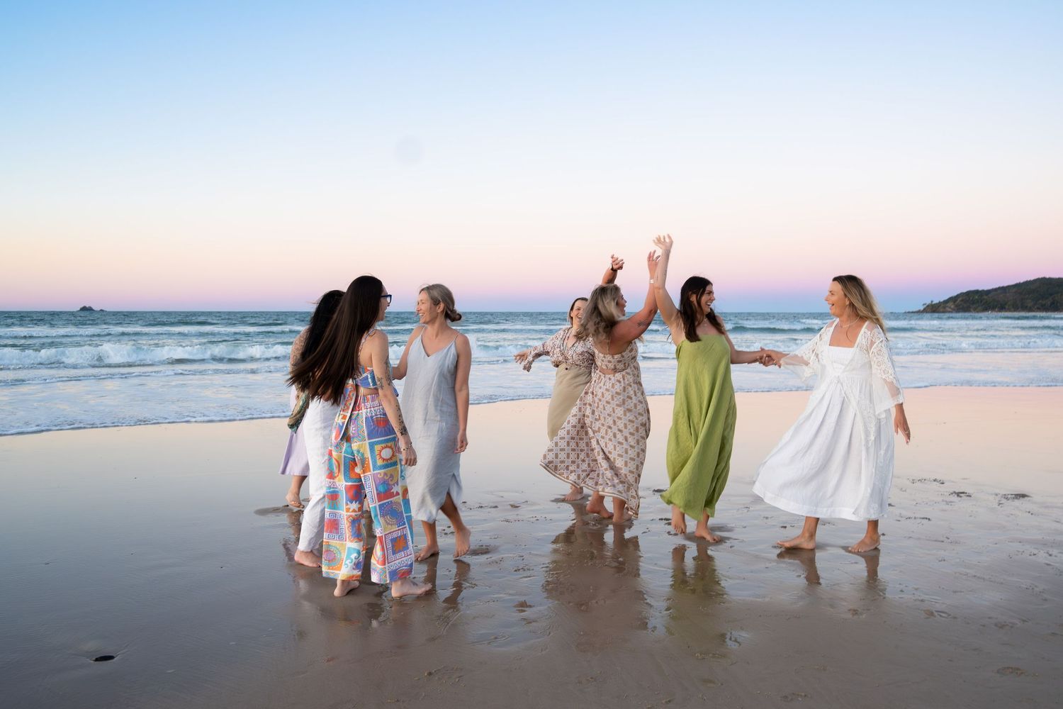 Group of women in flowing dresses dancing joyfully on a beach during a colorful sunset.