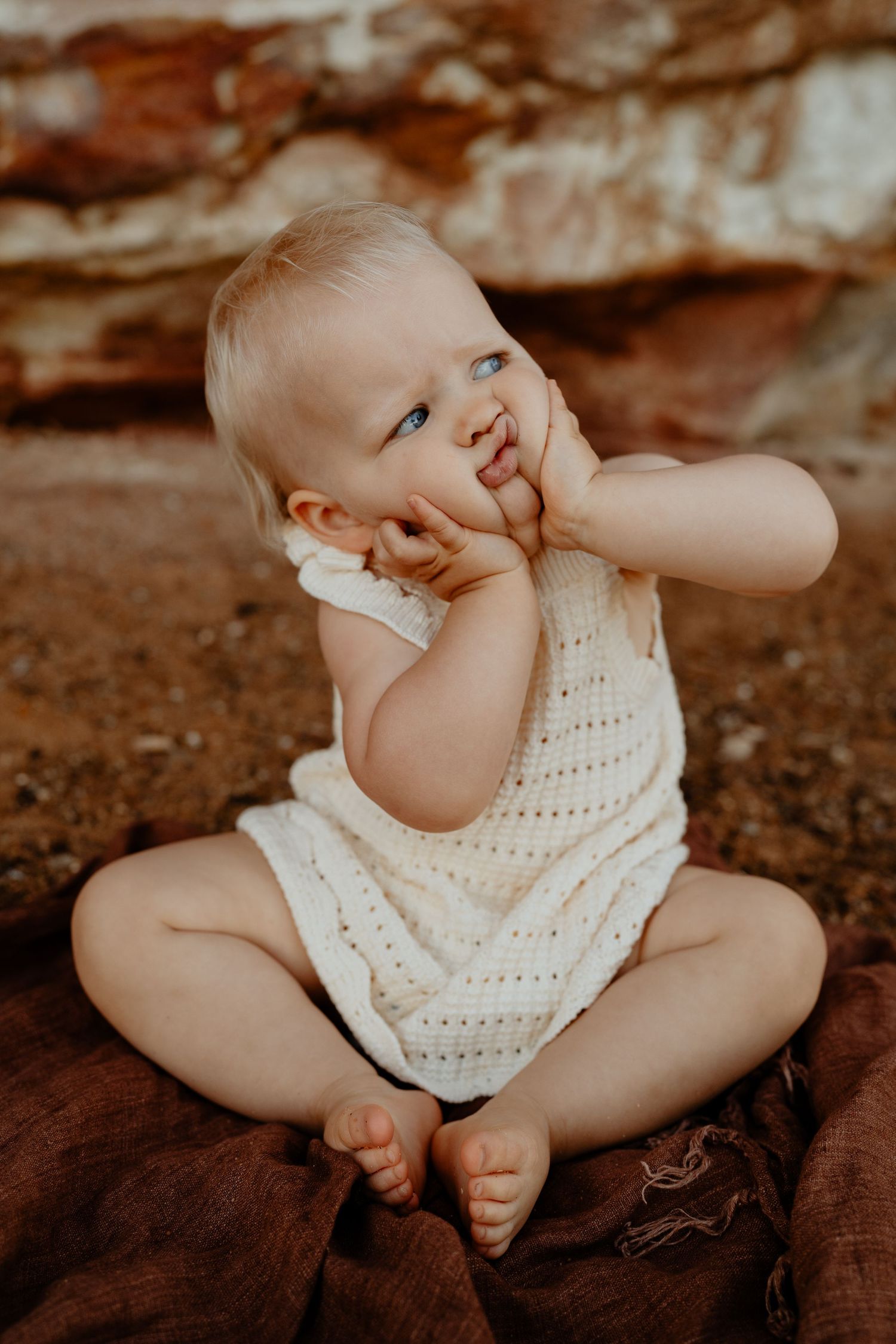 Cute baby in white knit romper sitting cross-legged on a blanket with fingers in mouth outdoors.
