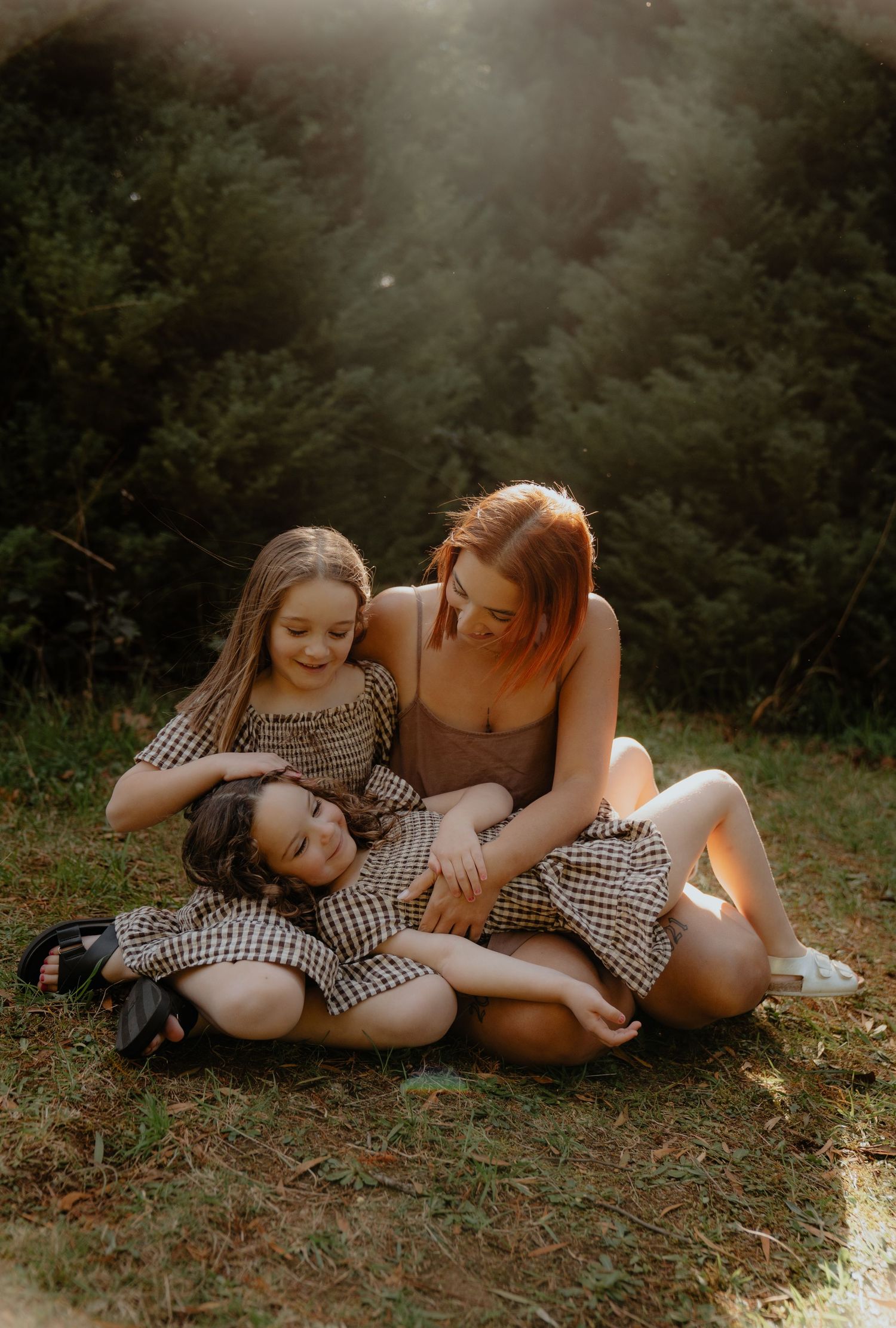 Mother sits on grass with two daughters in gingham dresses in golden sunlight.