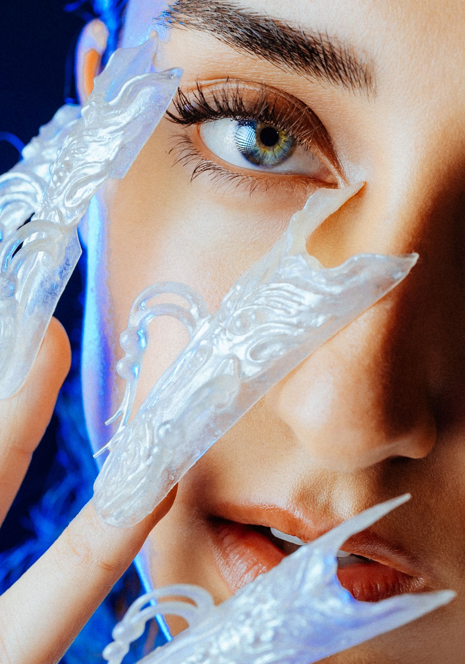 Close-up of a blue-eyed woman with ornate translucent decorative nail extensions near her face.