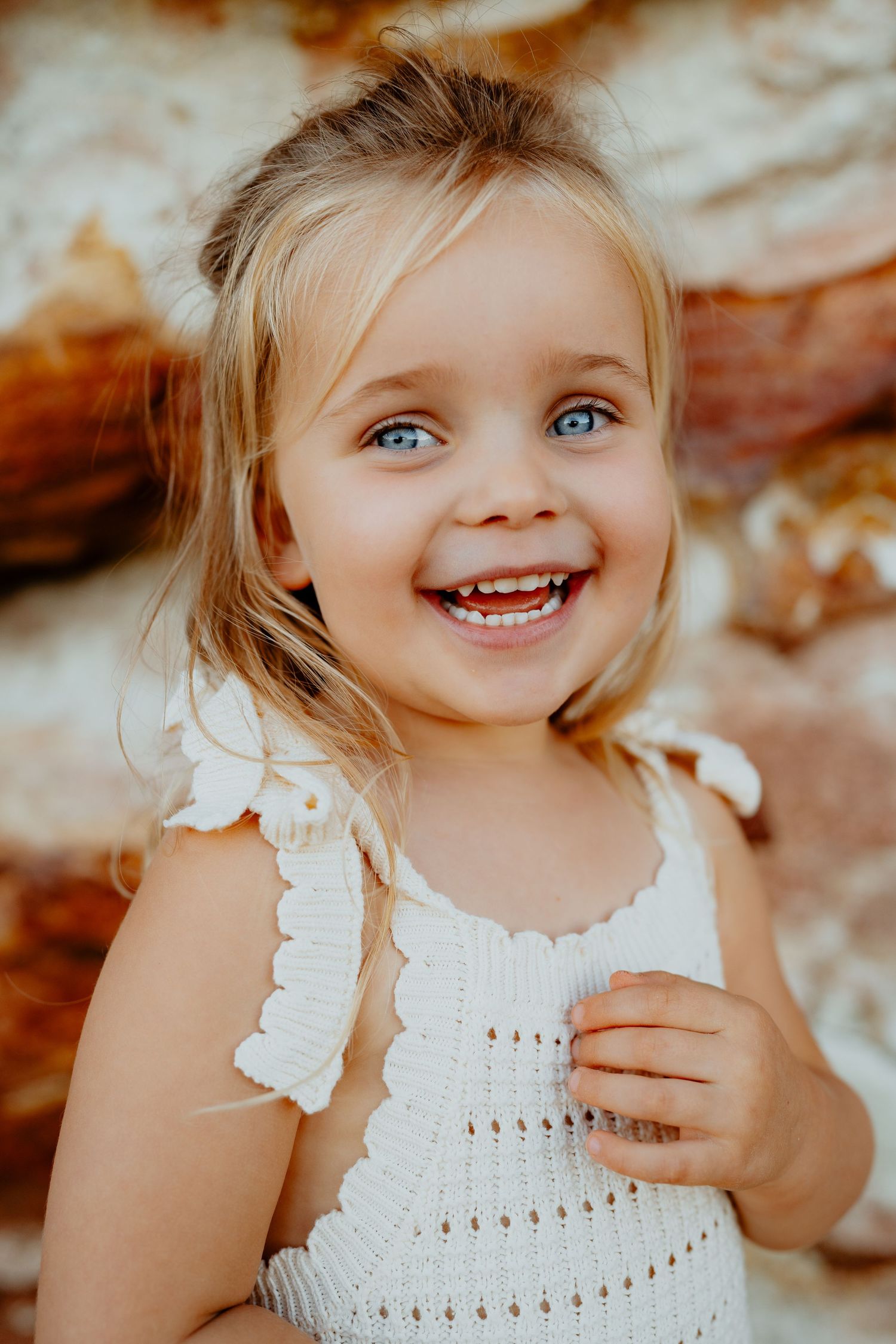 Adorable smiling blonde toddler girl with blue eyes wearing a white dress against a warm rocky background.