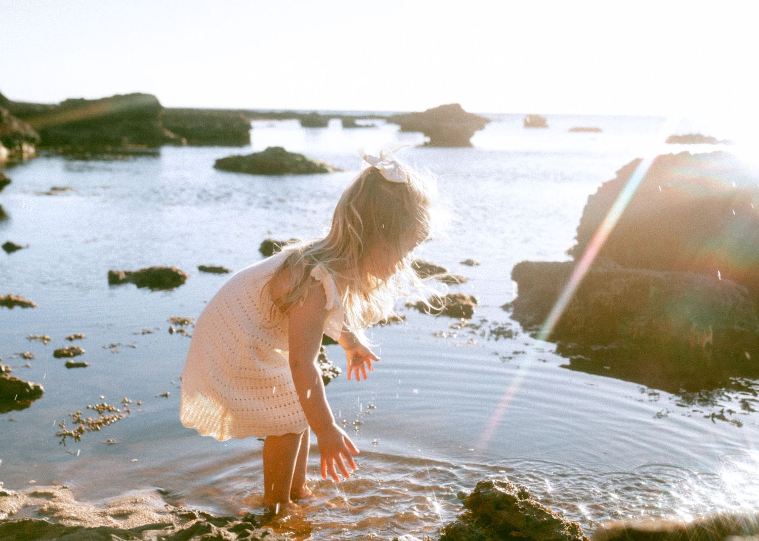 Young blonde girl in pink dress exploring rocky tide pools at the beach in sunlight.