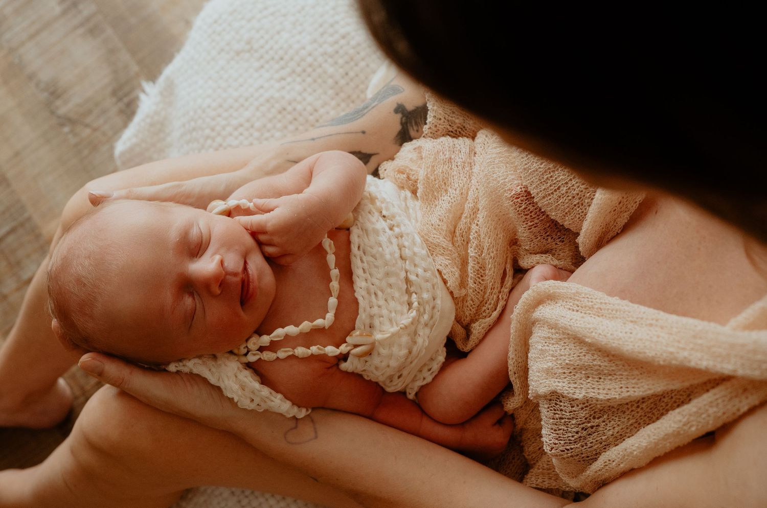 Mother cradling sleeping newborn wrapped in cream knit blanket with delicate pearl necklace.