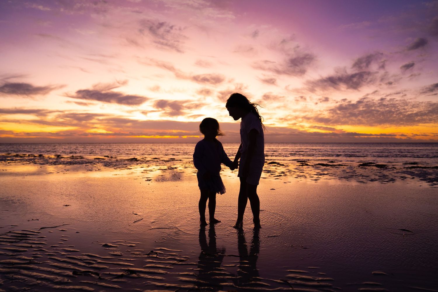 Silhouette of mother and child holding hands on a reflective beach during a vibrant purple sunset.