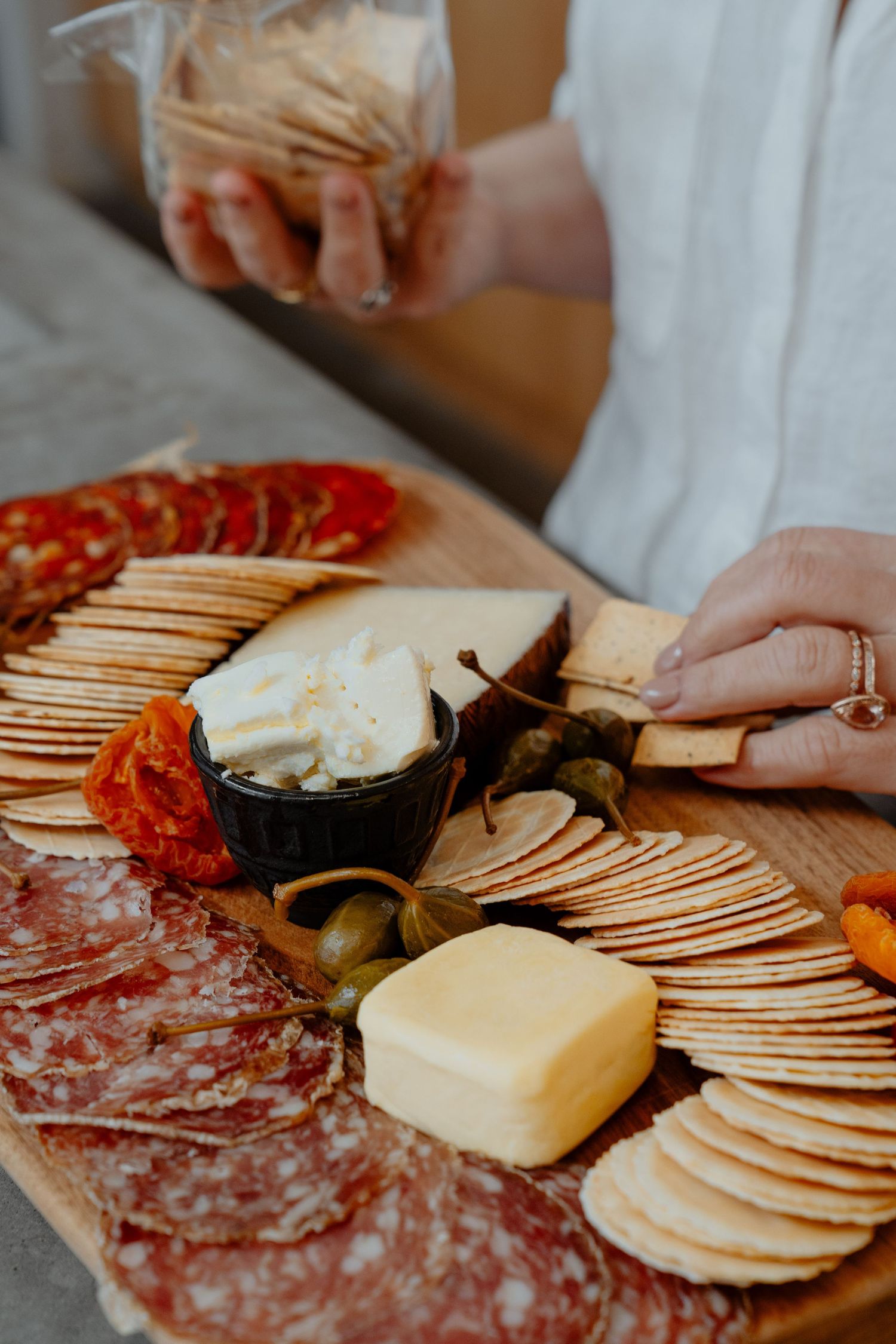 Woman arranging a gourmet charcuterie board with salami, cheese, crackers, and olives on a wooden platter.