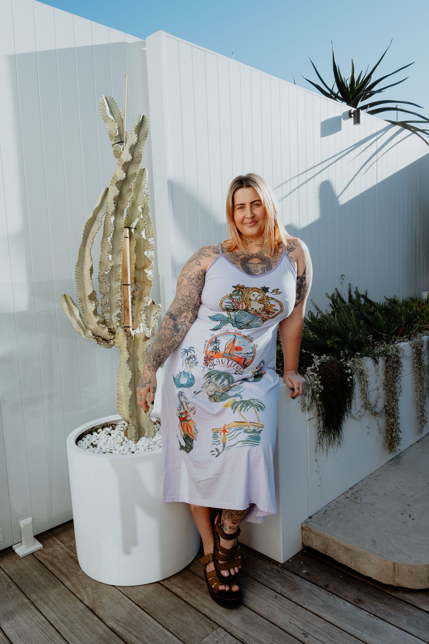 Tattooed woman in a graphic midi dress posing beside a gold cactus sculpture on a white outdoor patio.