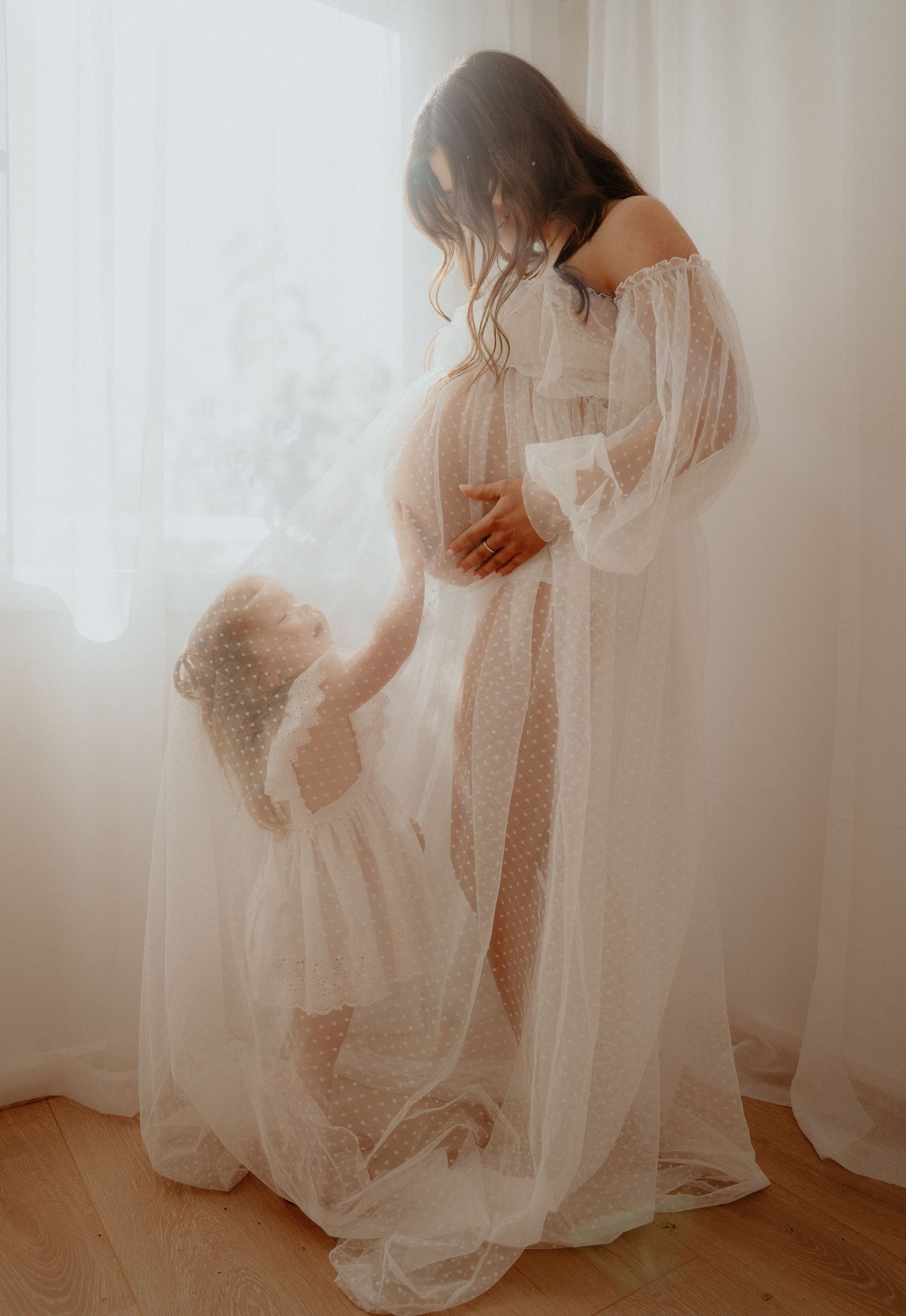 A little girl in a white dress lovingly touches a pregnant womans belly as they stand together by a bright window.