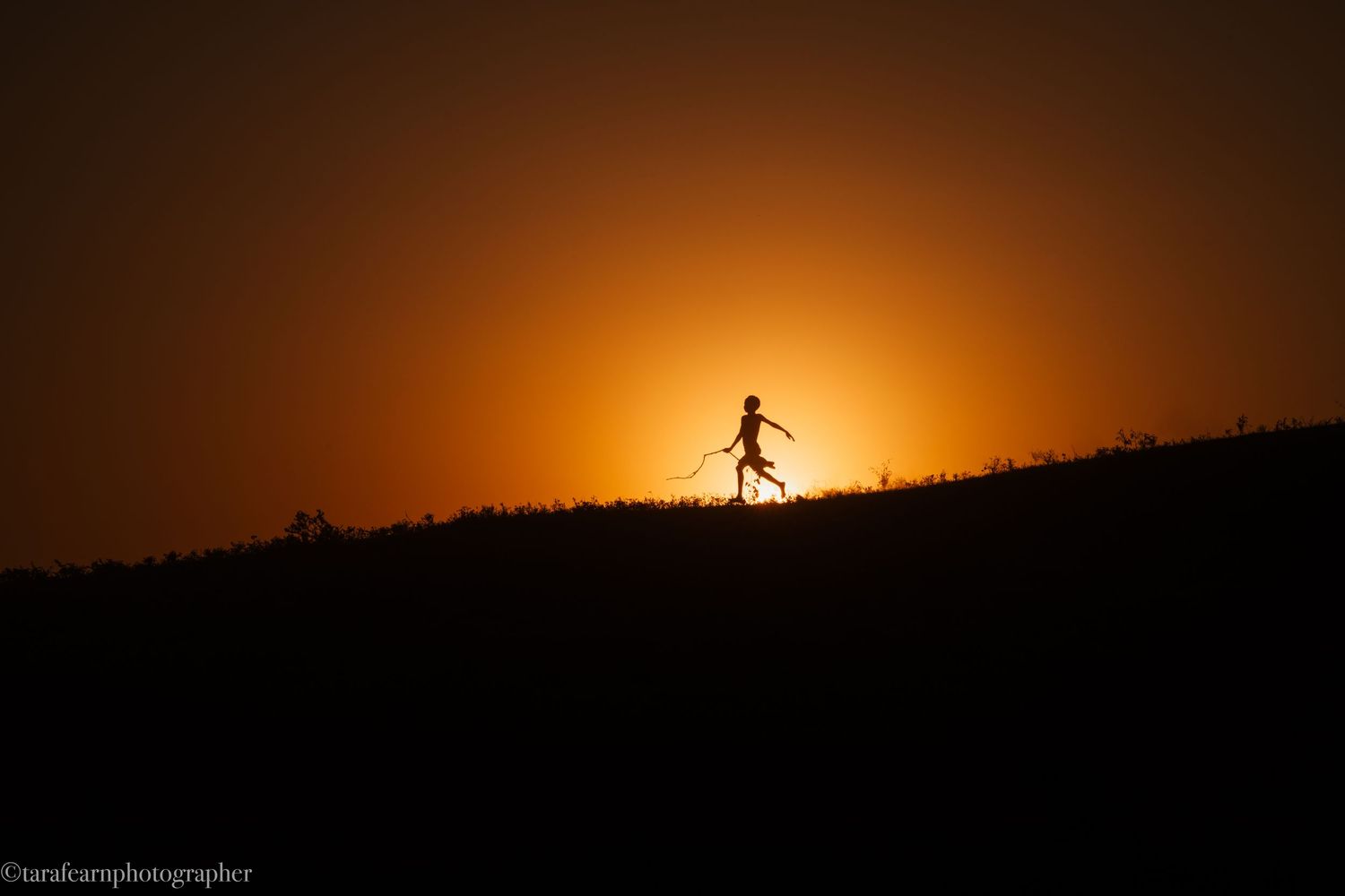 Silhouette of a child running along a hilltop against a glowing orange sunset sky.