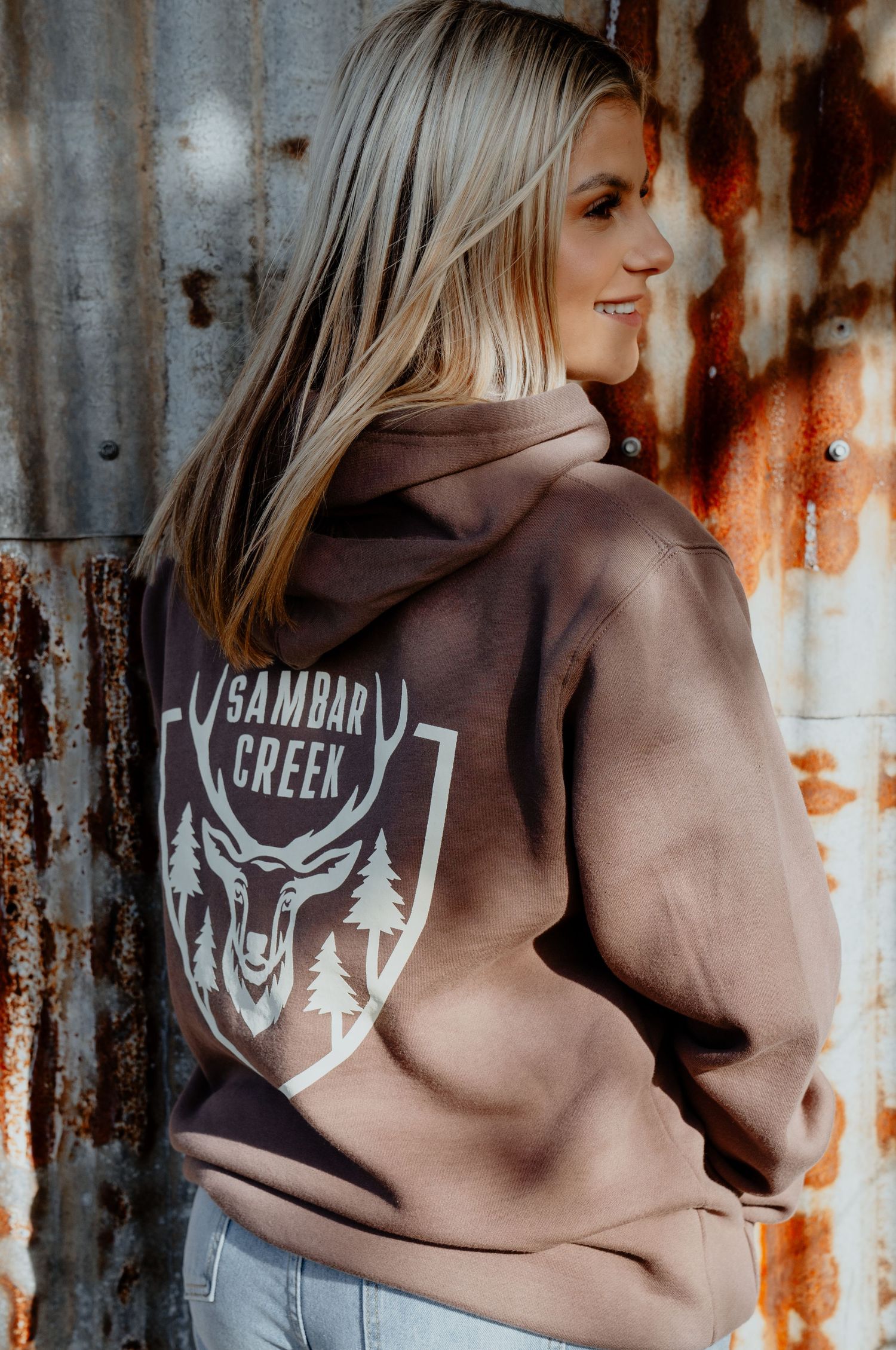 Blonde woman wearing brown Sambar Creek branded hoodie with deer shield logo, against rustic metal wall.