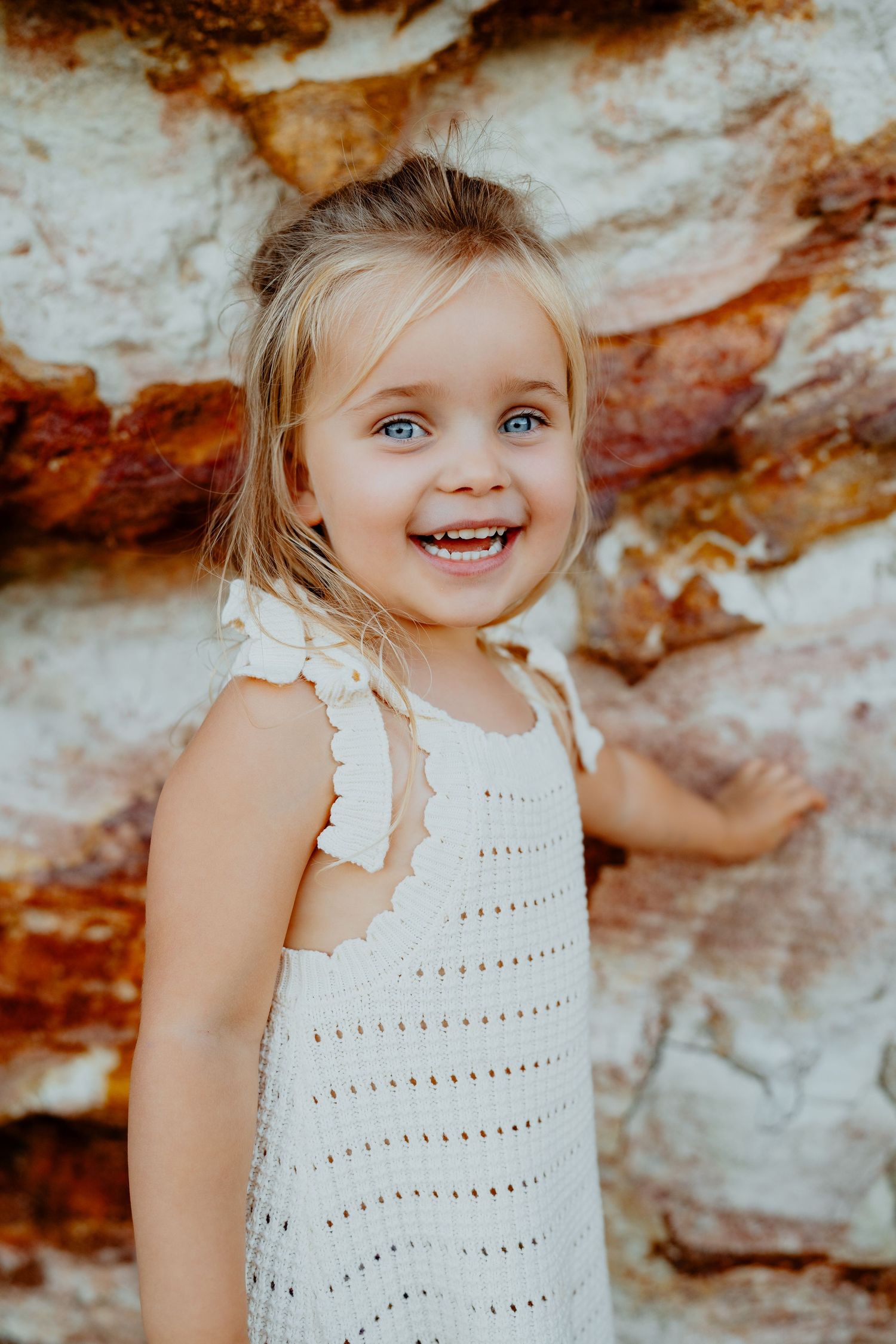 Adorable blonde blue-eyed little girl in white dress laughing joyfully against a rustic rock wall.