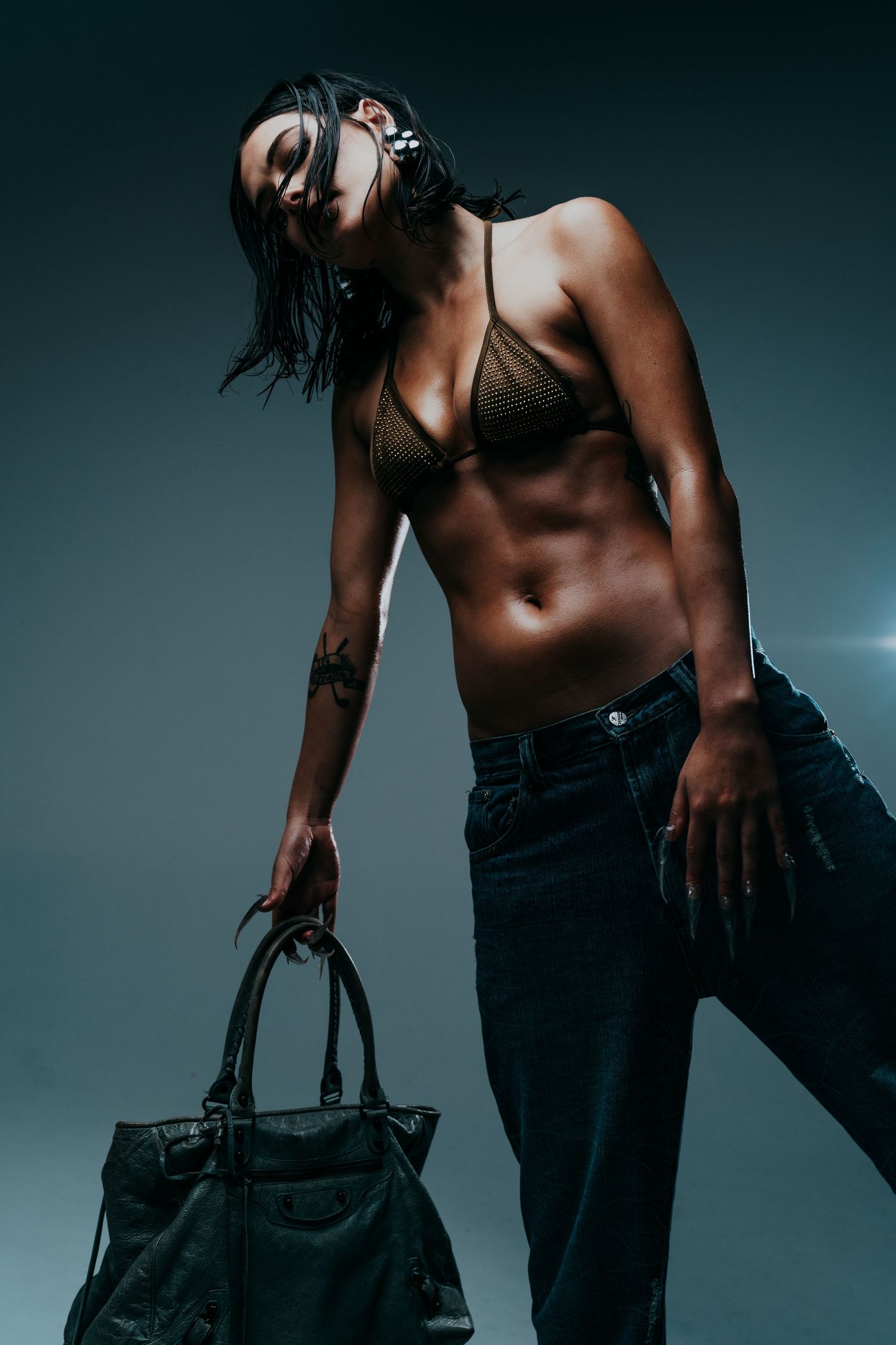 Woman in olive bikini top and jeans holding leather bag in dark studio setting.