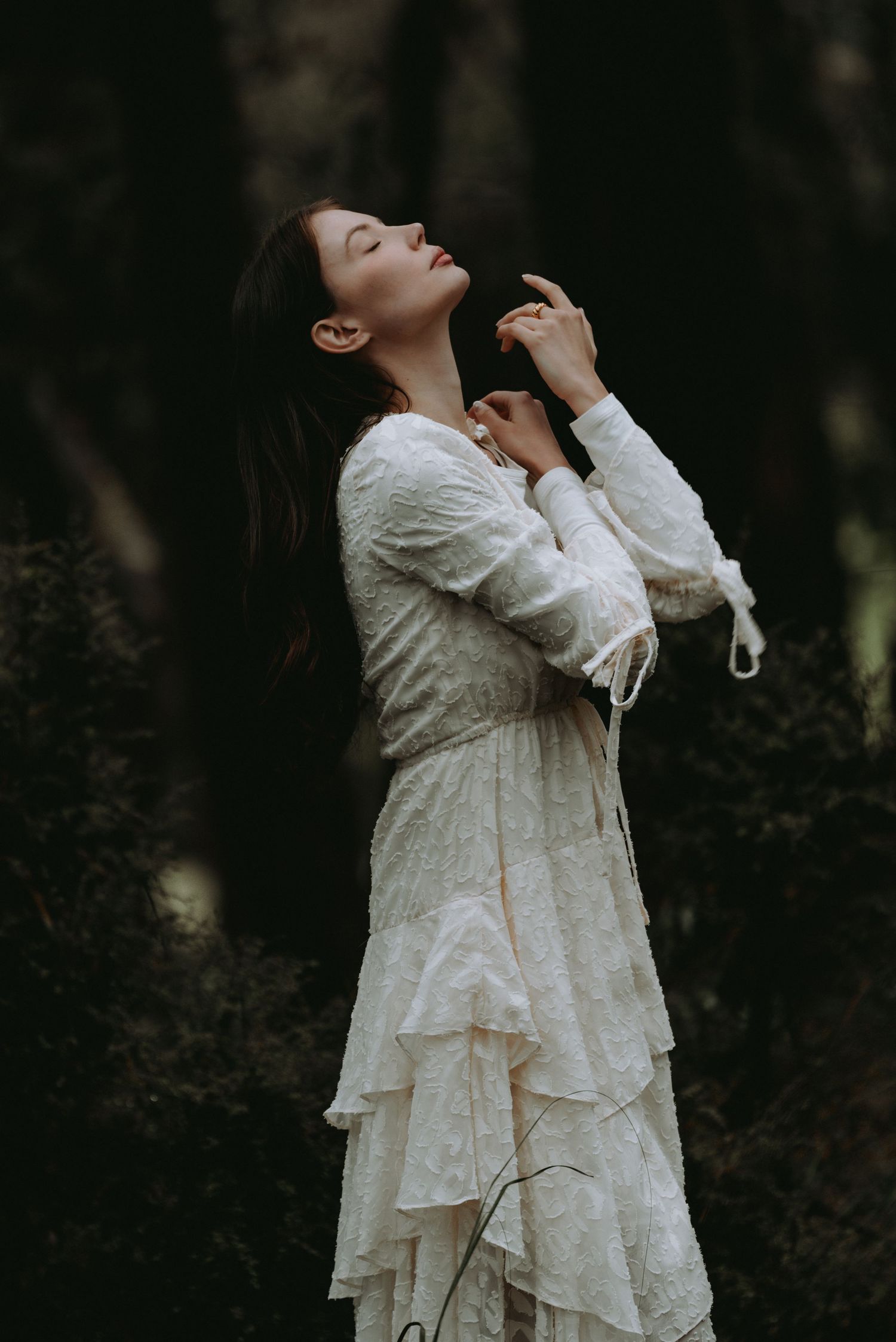 Woman in white ruffled lace dress standing in dark forest with eyes closed, head tilted back.