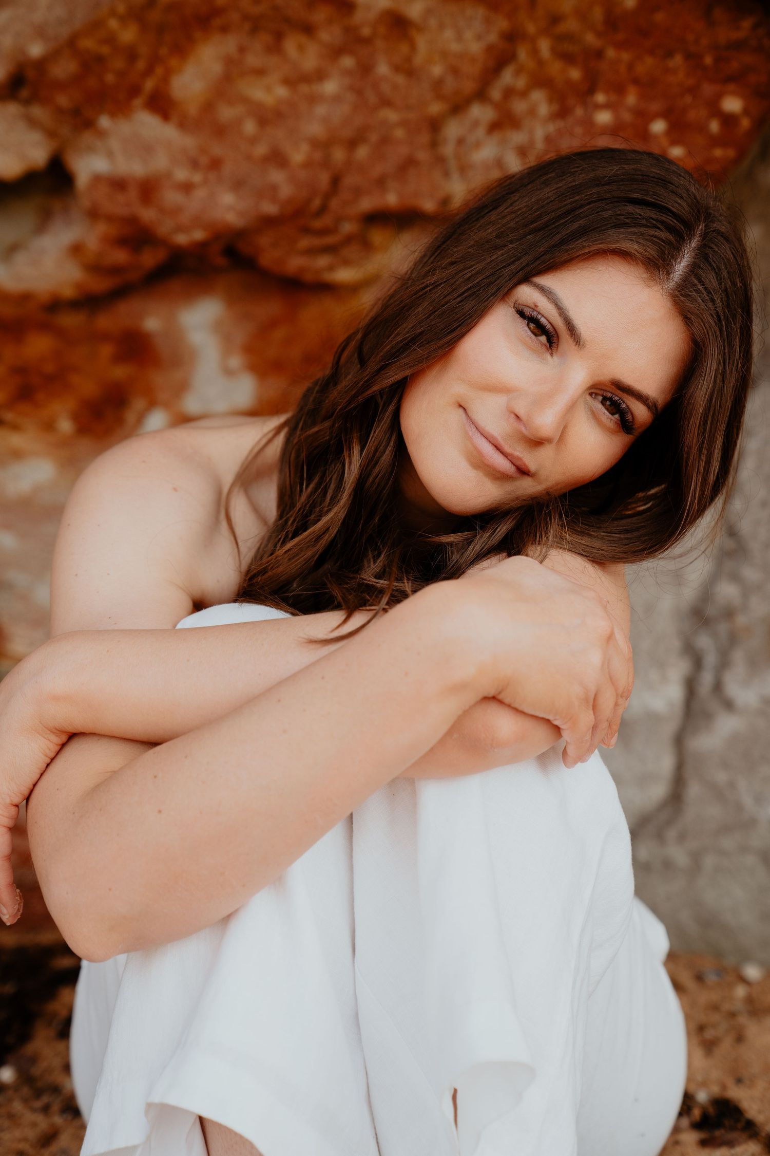 Brunette woman in white outfit sitting against a red rock wall smiling softly.