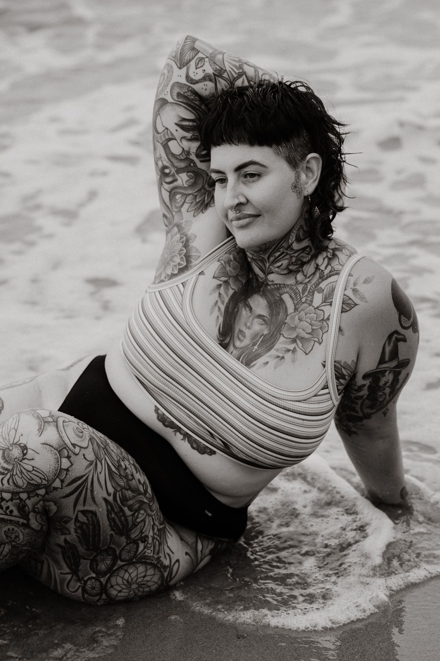 Black and white photo of heavily tattooed woman in wrapped top sitting in ocean waves on beach.