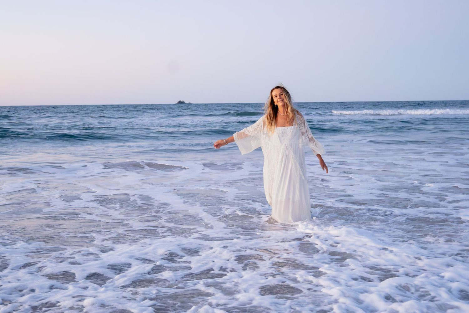 Woman in a flowing white maxi dress standing gracefully in ocean waves at the beach.