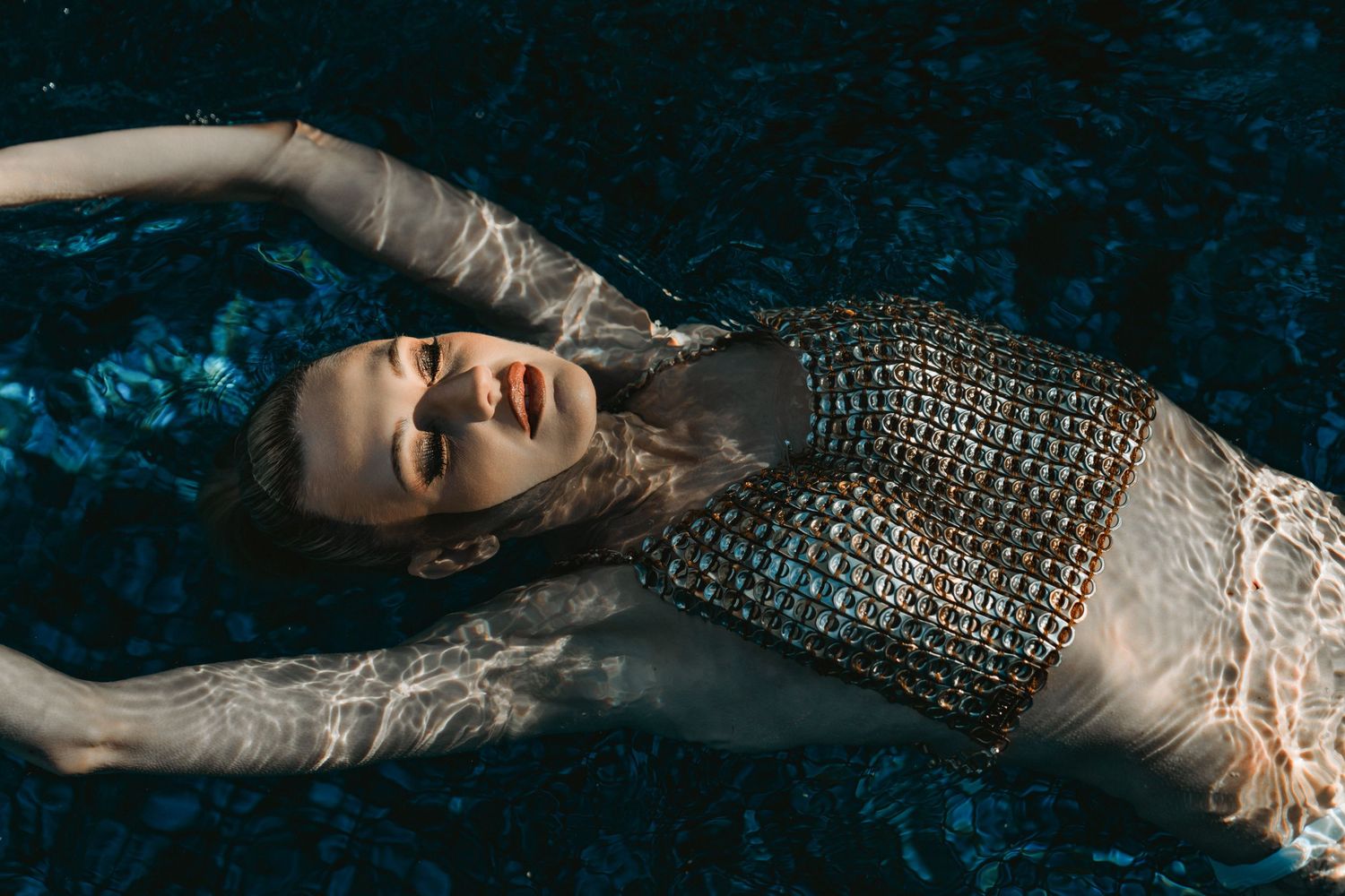 Woman in metallic chainmail top floating in dark shimmering pool water with eyes closed peacefully.