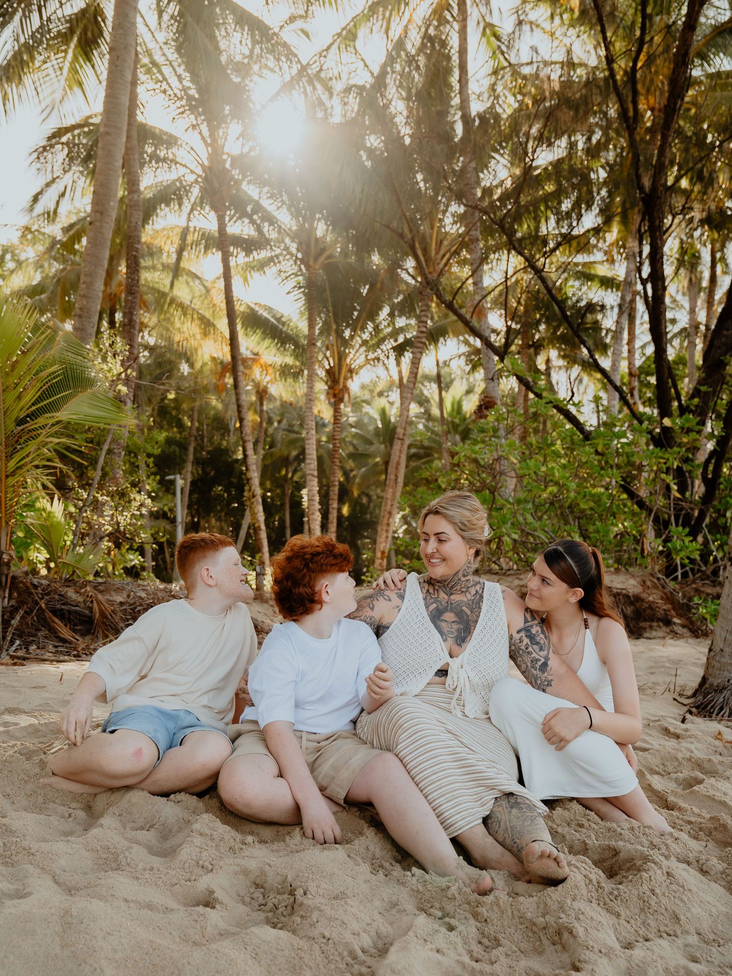 Family of four sitting together on a sandy beach surrounded by tropical palm trees.