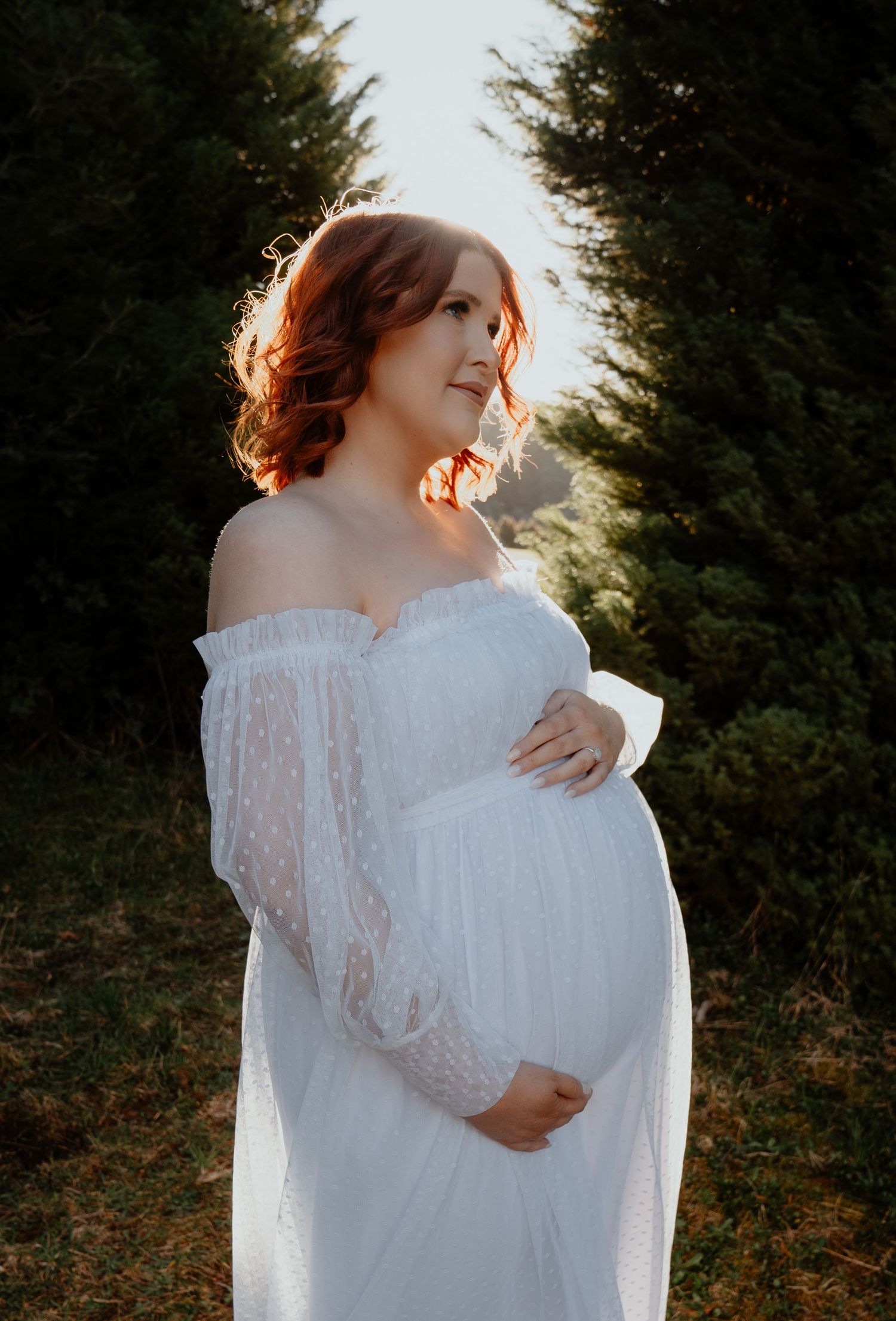 Pregnant woman in white off-shoulder lace dress cradling belly among evergreen trees.
