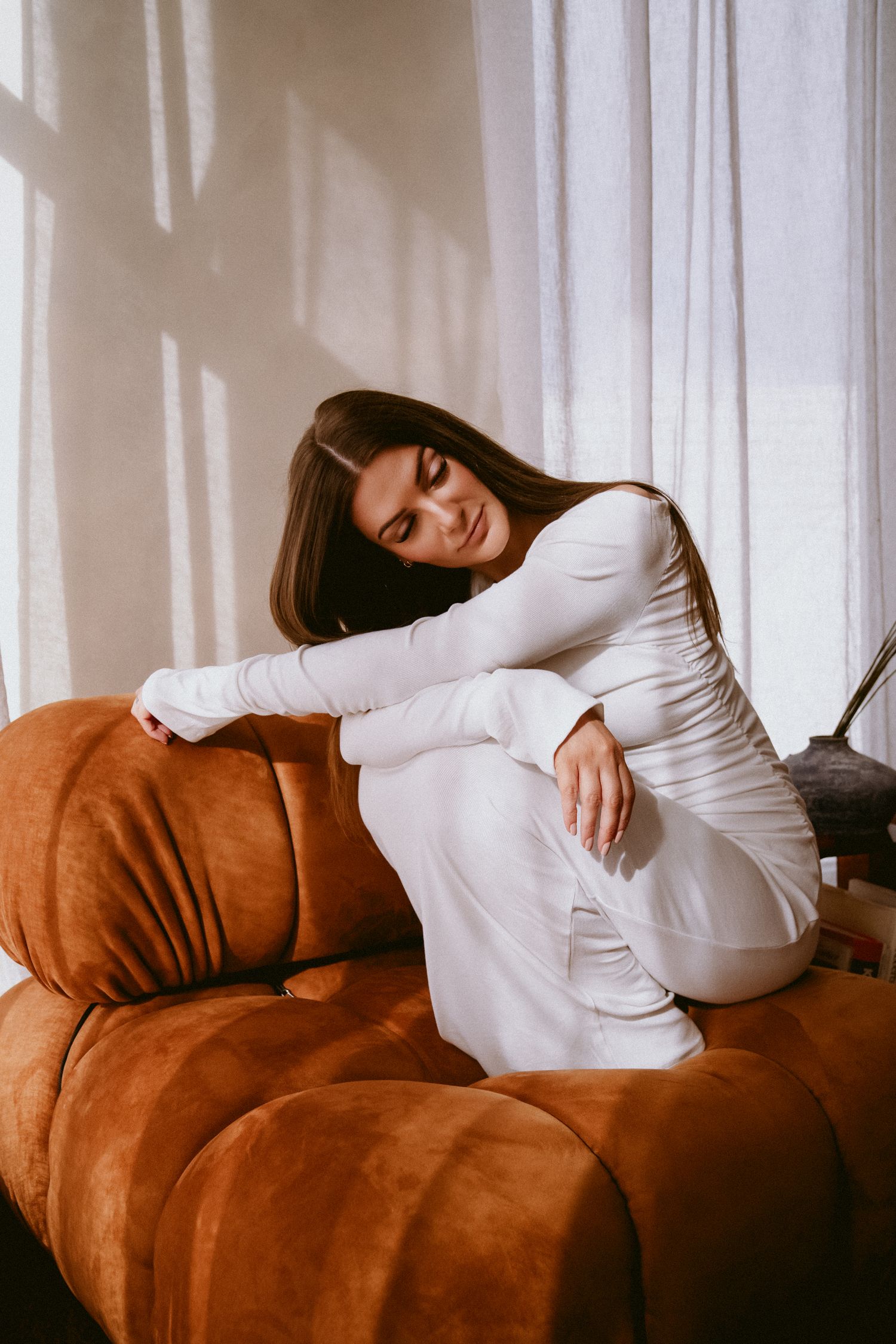 Brunette woman in white outfit relaxing on an orange velvet sofa near sheer curtains in soft light.