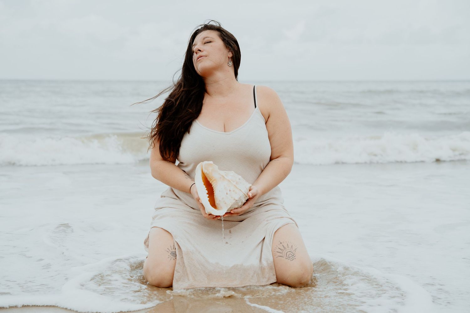 Curvy woman in wet beige slip dress kneeling in ocean waves holding a large conch shell.