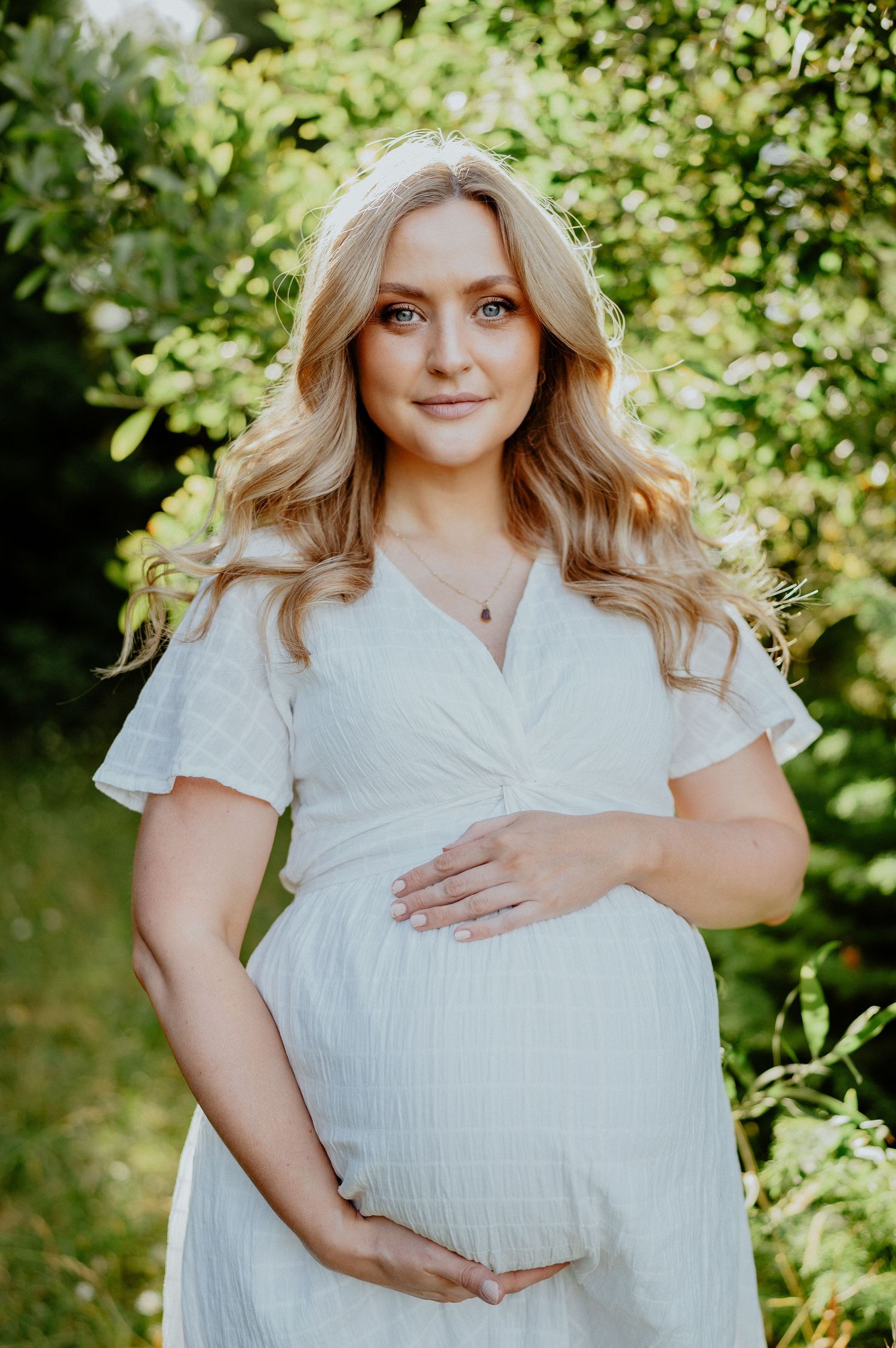 Pregnant woman in white dress cradling baby bump in a lush green garden.