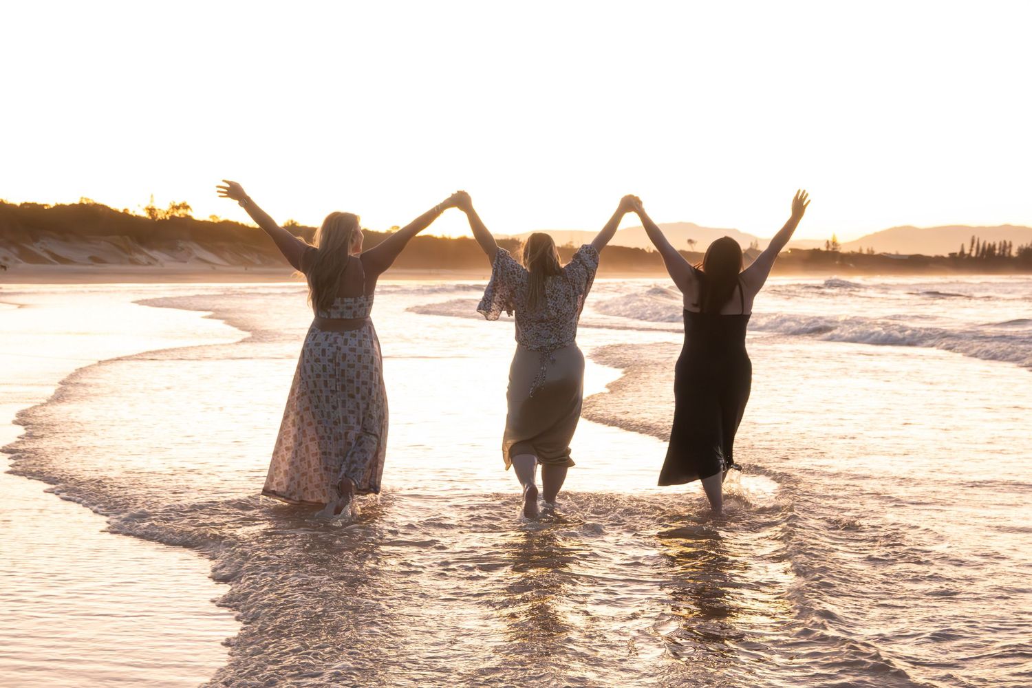 Three women holding hands raised toward the sky while wading in golden sunset beach waves.