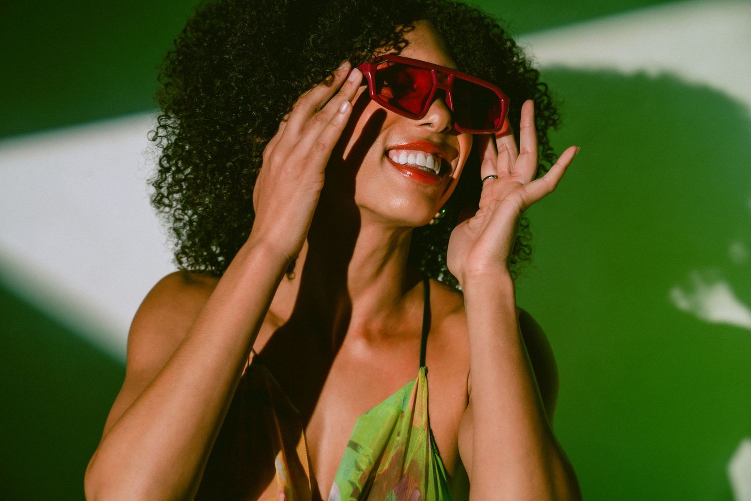 Smiling woman with curly hair adjusting red oversized sunglasses wearing green floral dress.