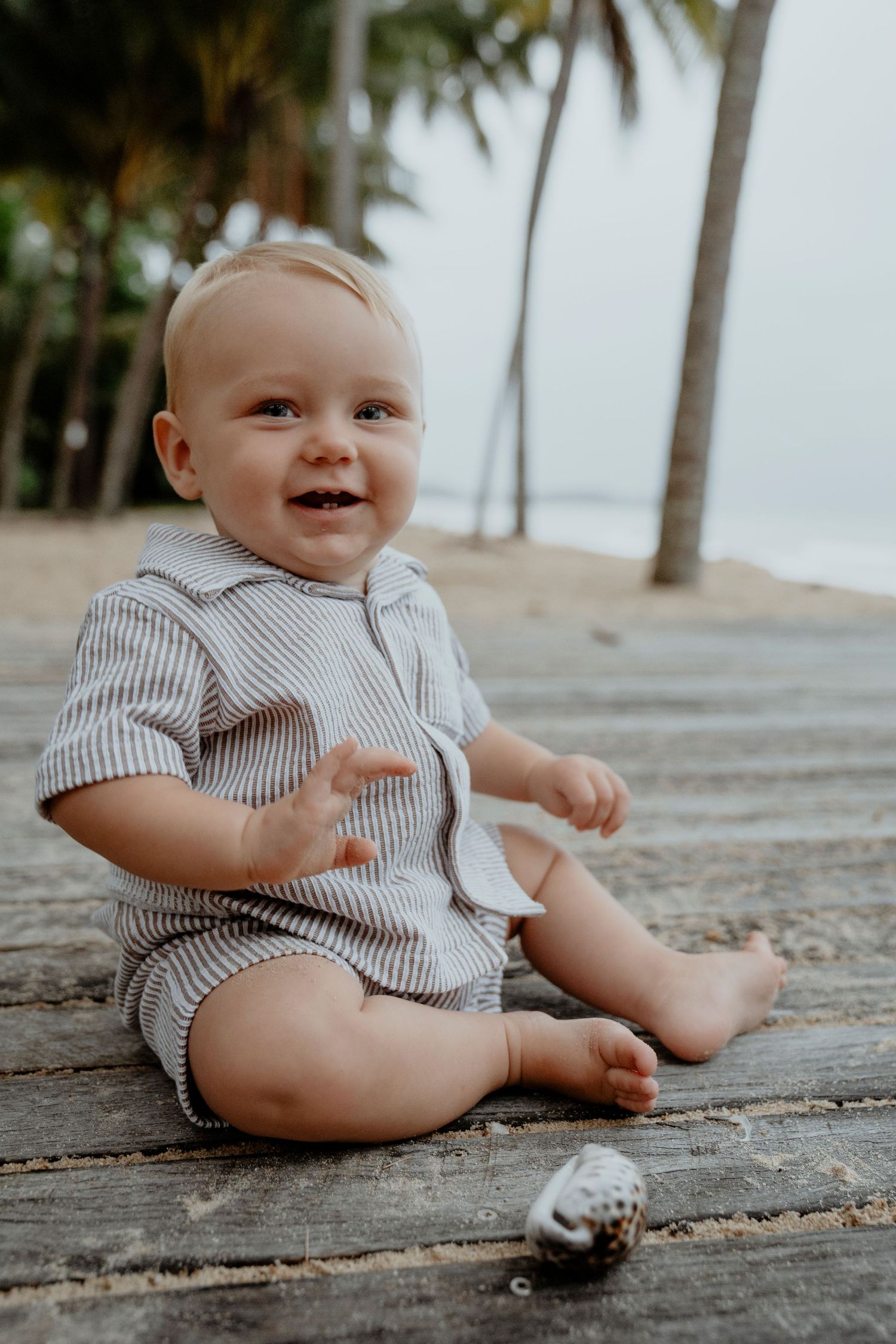 Happy baby in striped romper sitting on wooden deck at tropical beach with palm trees and seashell nearby.