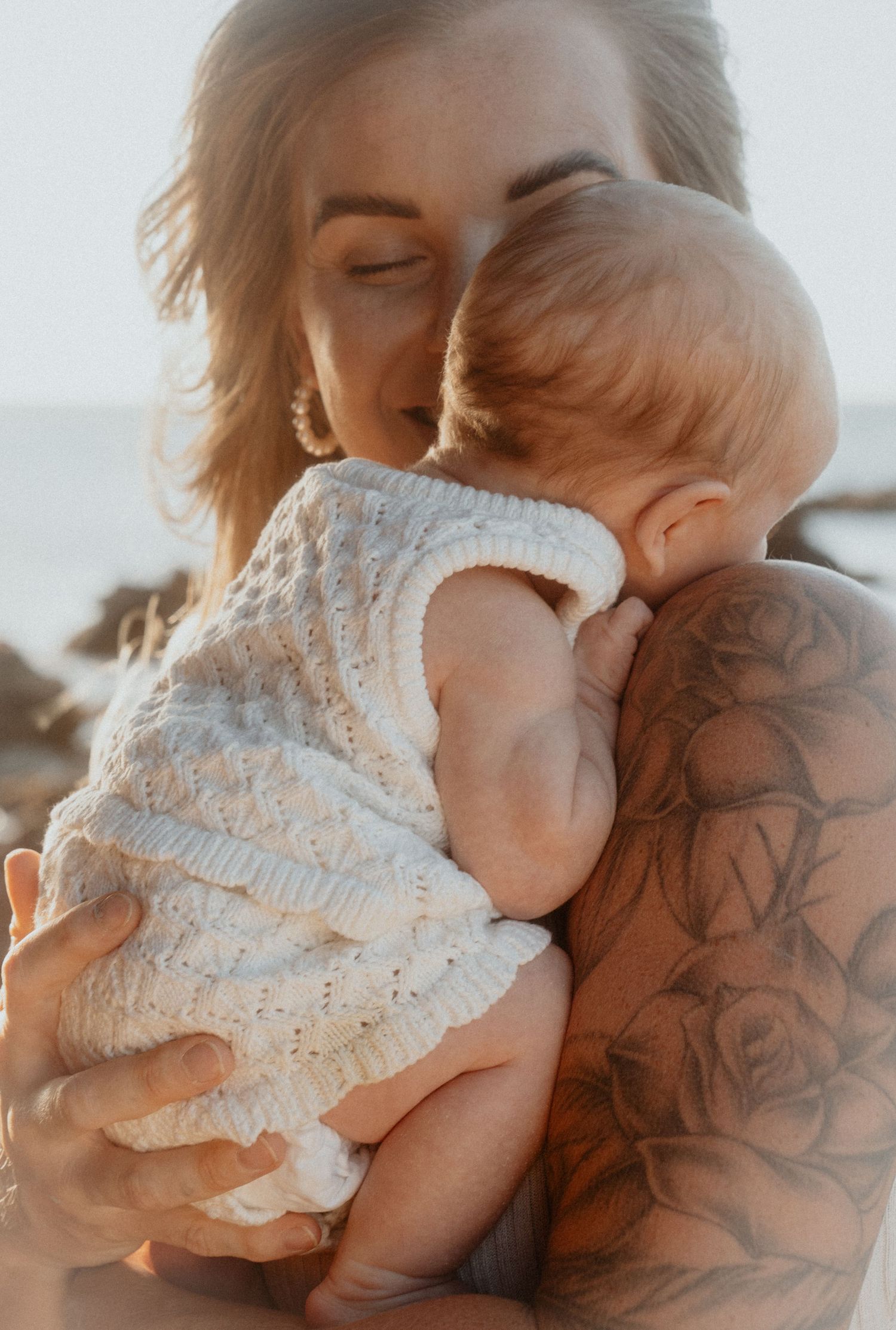 Tattooed mother gently kissing her newborn baby on the head while cradling them at the beach.
