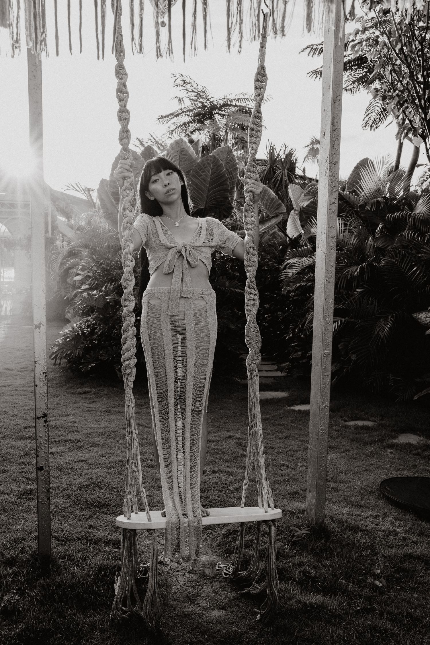 Young woman standing on a macrame swing surrounded by lush tropical greenery in a black and white photo.
