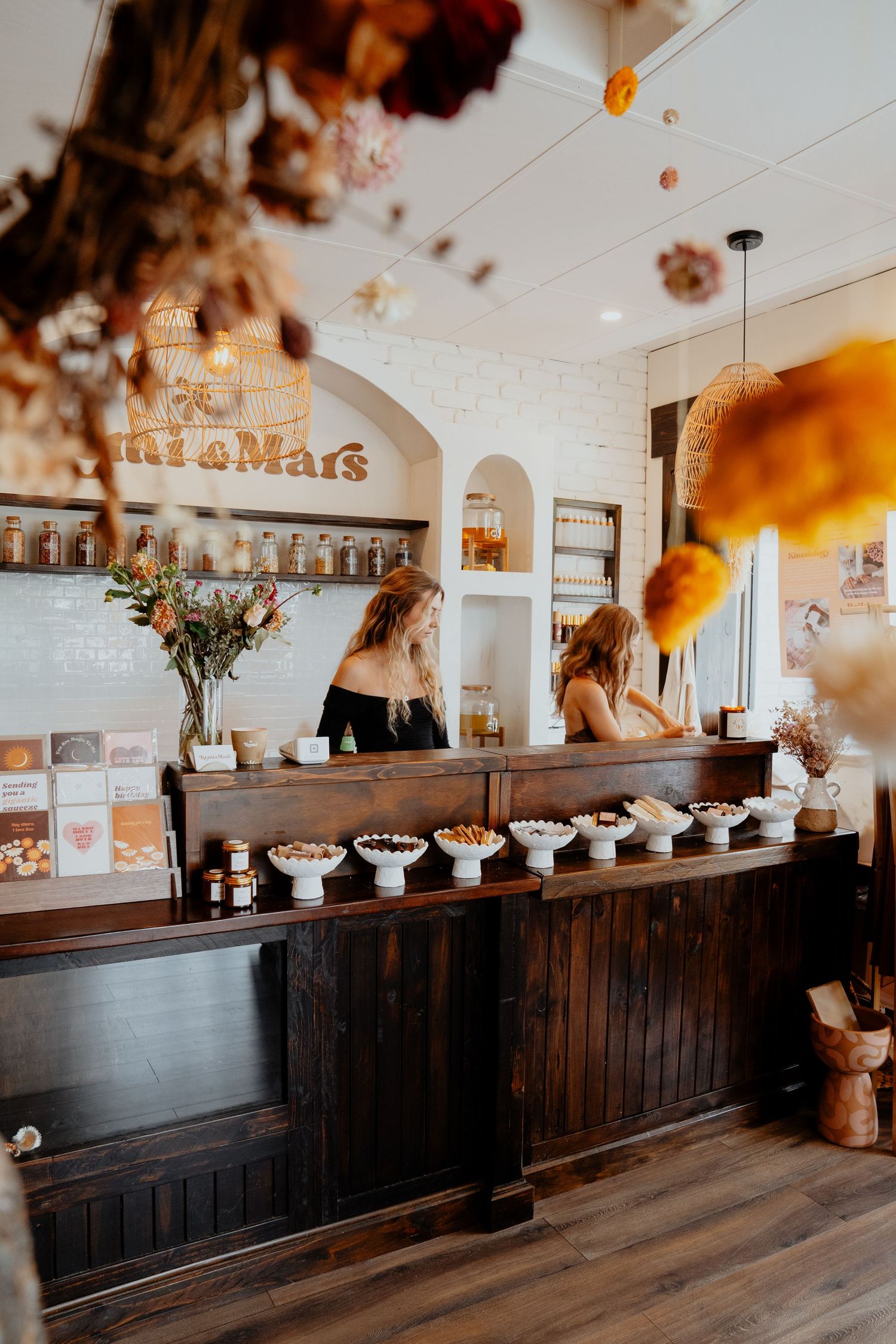 Two women working behind a dark wooden counter at Remi and Mars boutique shop with warm boho decor.