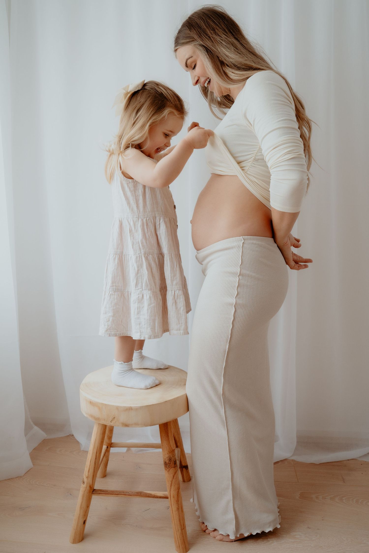 A smiling pregnant woman in a white crop top stands beside a little girl on a wooden stool in a bright studio.