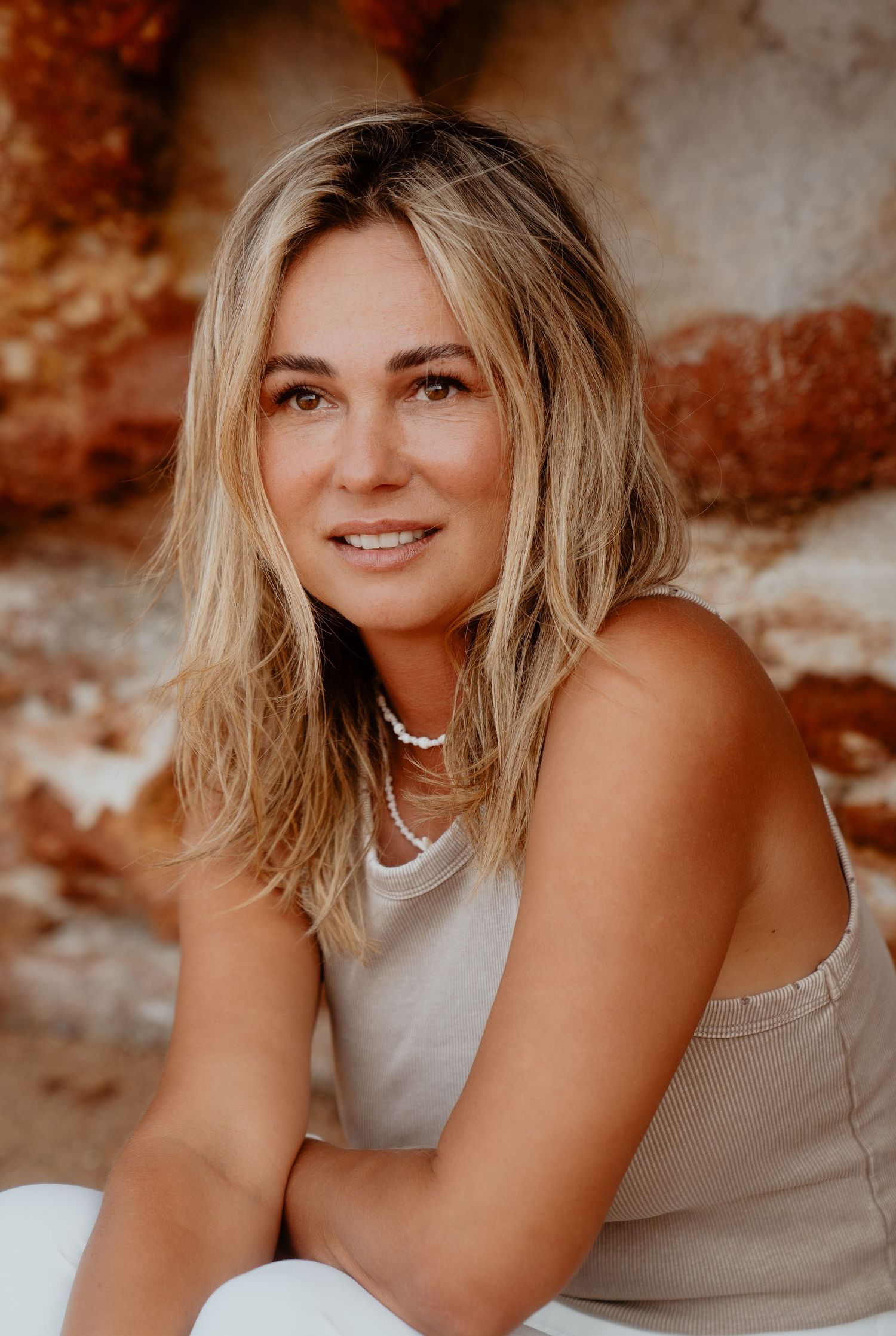 Blonde woman in a beige tank top with a pearl necklace sitting near rocky terrain.