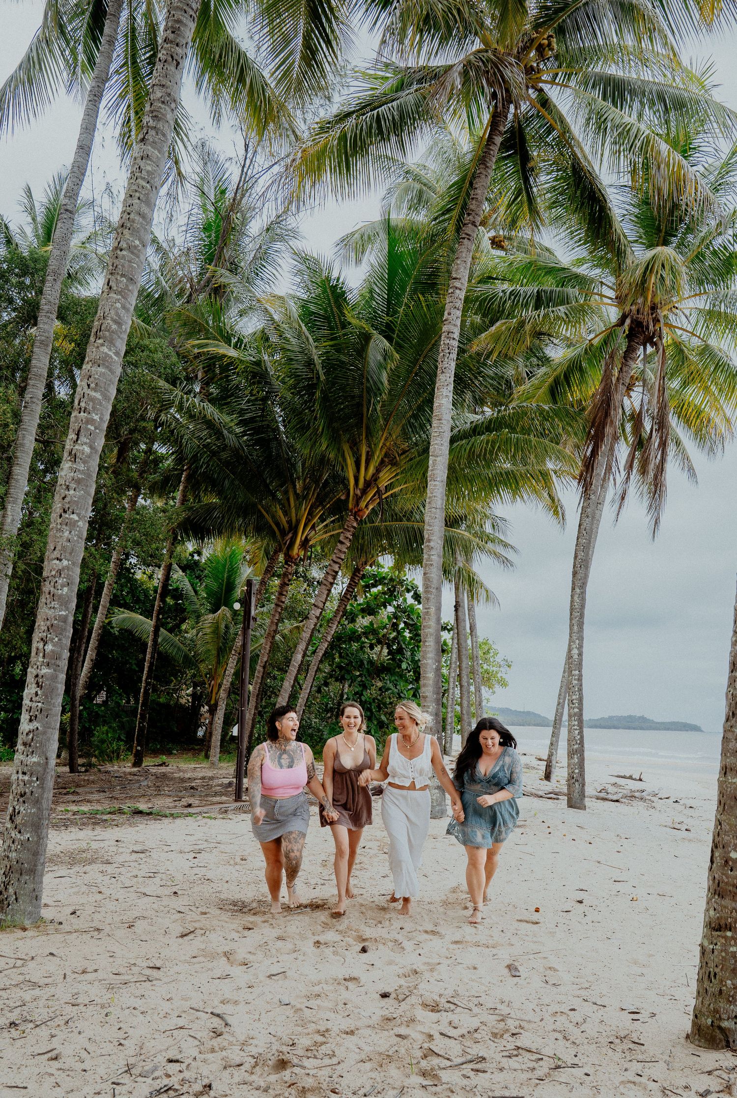 Four women walking together on a tropical white sand beach lined with tall palm trees.