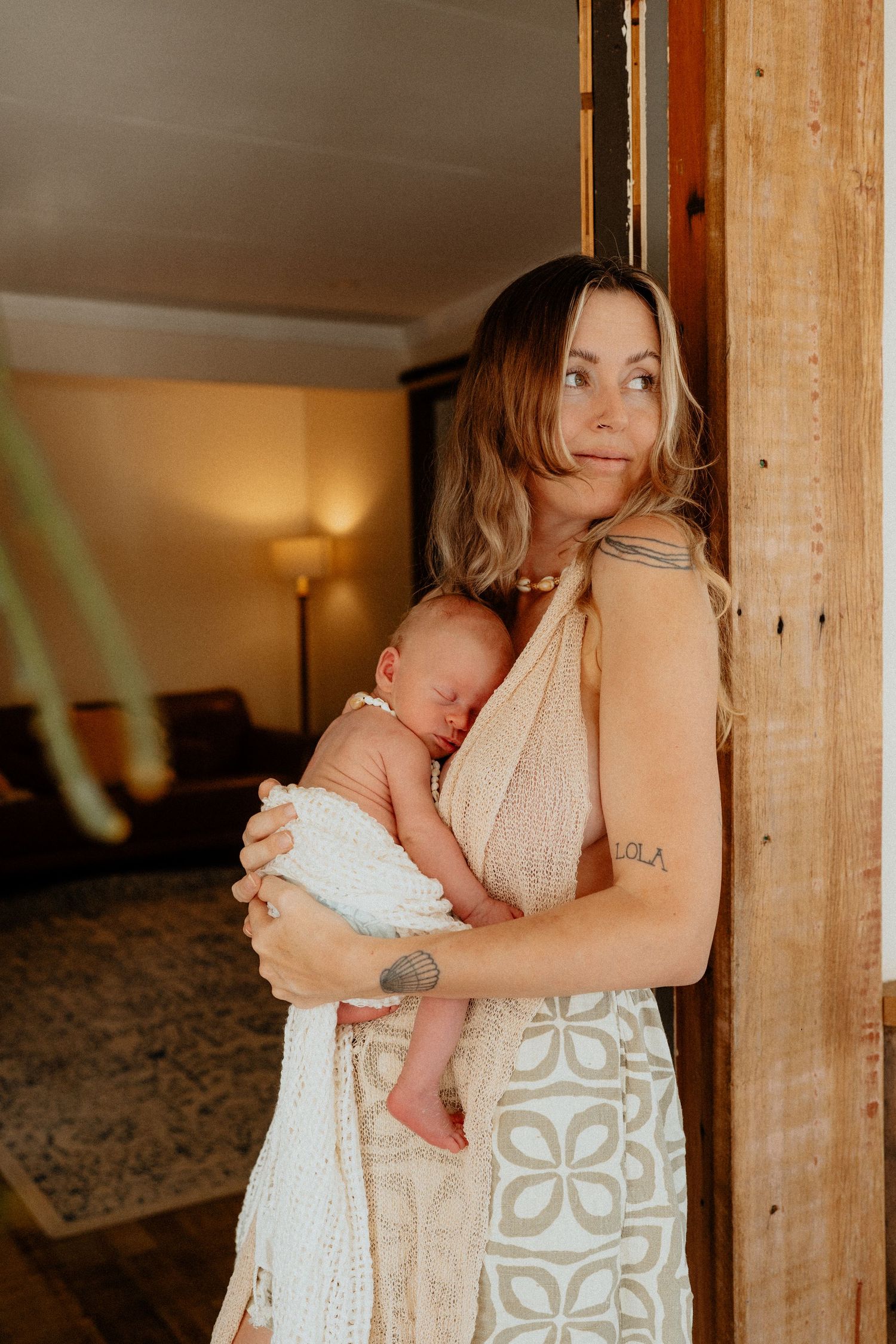 Mother in boho dress holding swaddled newborn baby while leaning against rustic wooden doorframe.