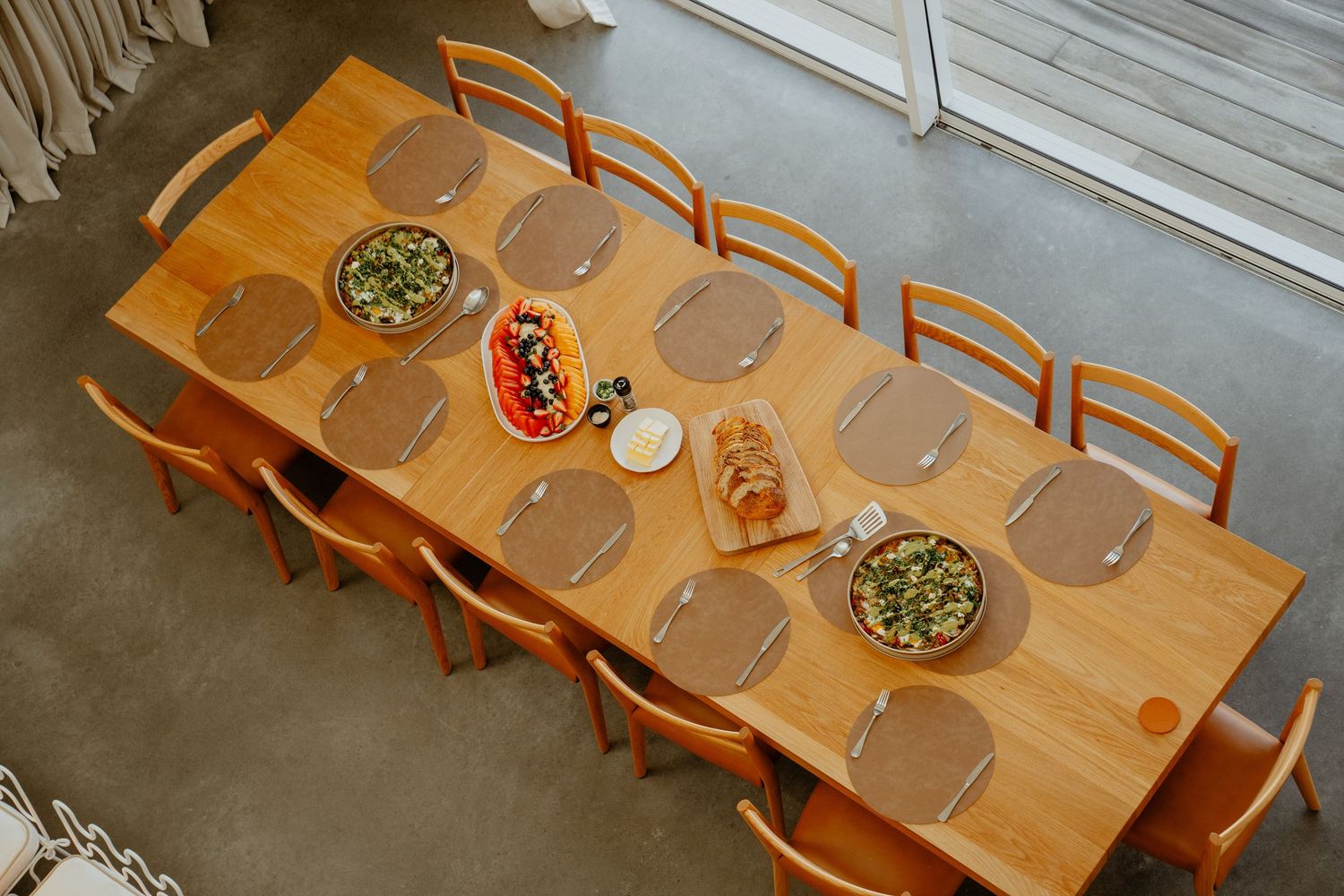 Aerial view of a wooden dining table set for a group meal with salads and bread in a bright room.