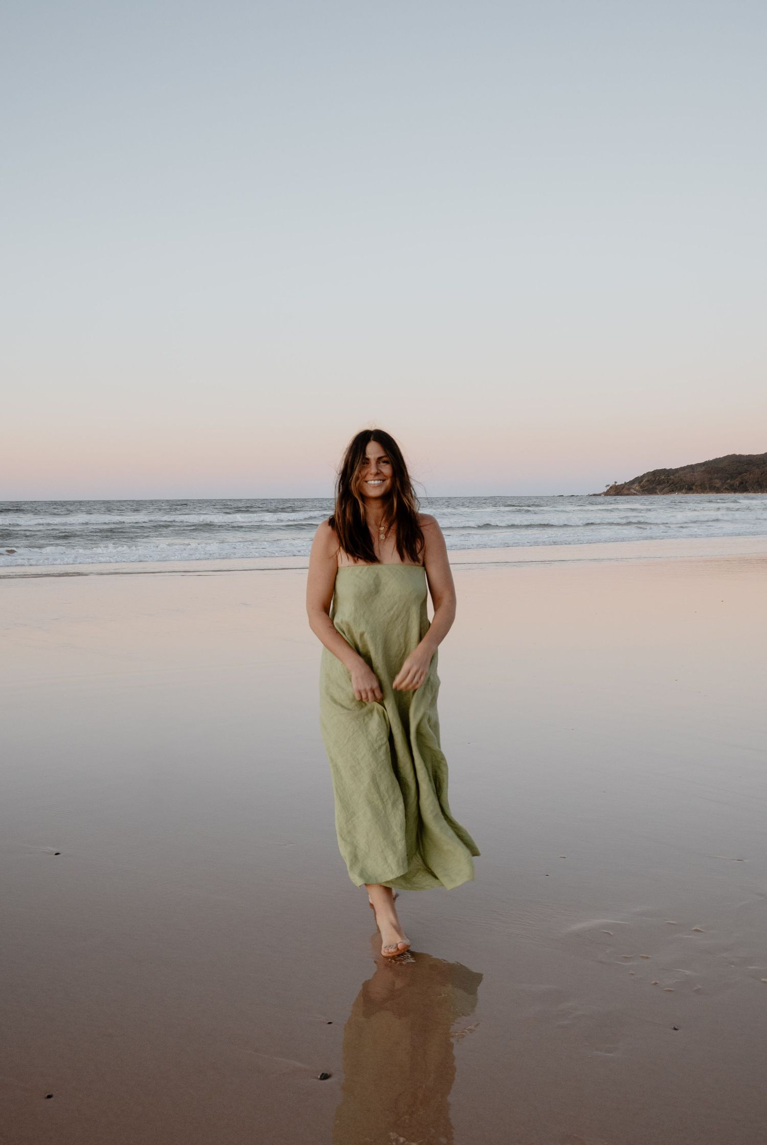 Woman in a flowing green linen maxi dress walking barefoot on a beach at sunset.