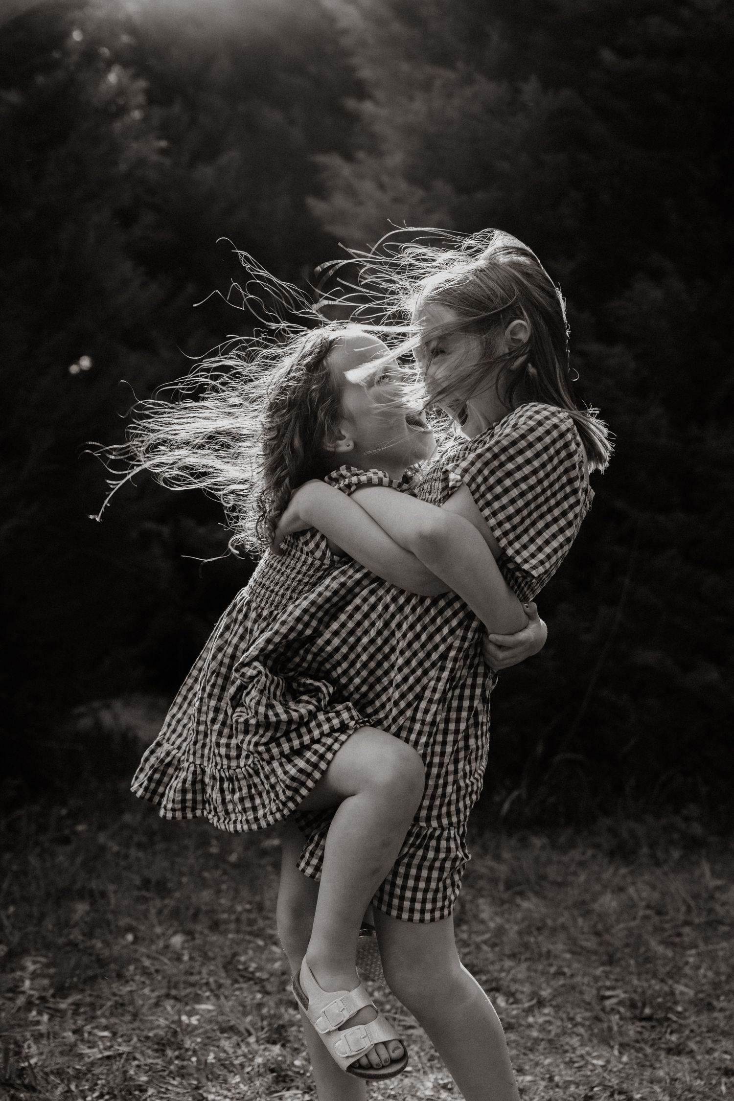 Black and white photo of two girls in gingham dresses embracing in the wind.