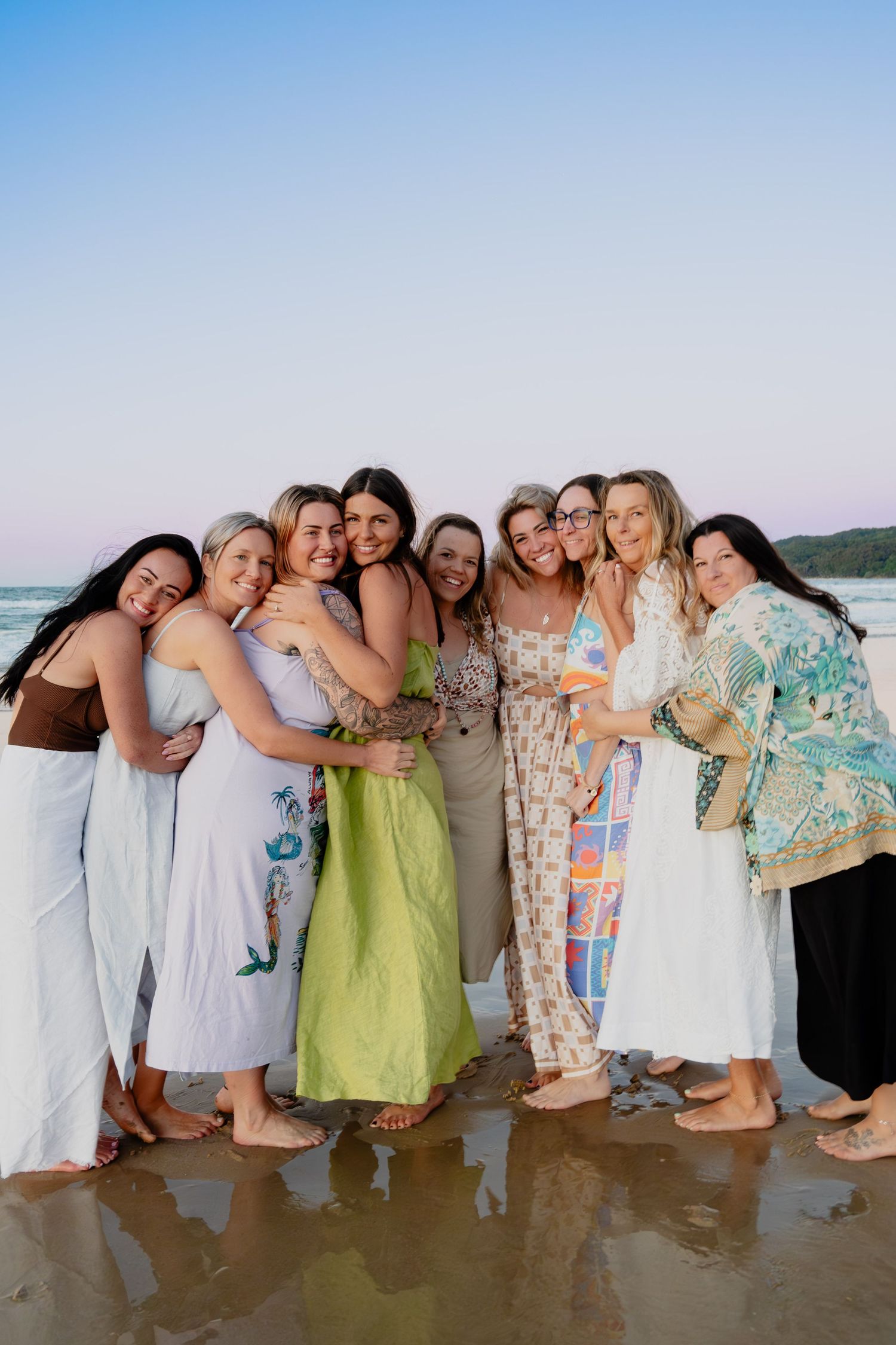 Group of ten smiling women hugging together on a reflective wet beach at twilight.