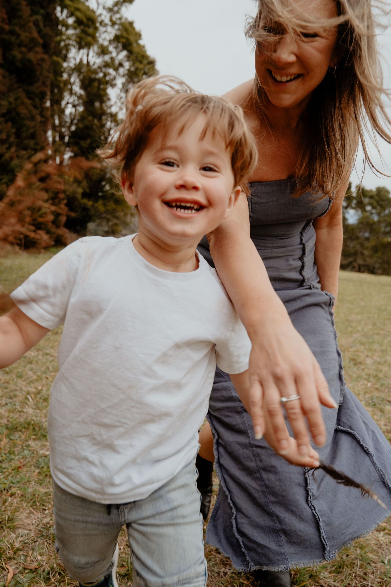 Happy toddler boy in white shirt laughing with smiling mother in a green park.
