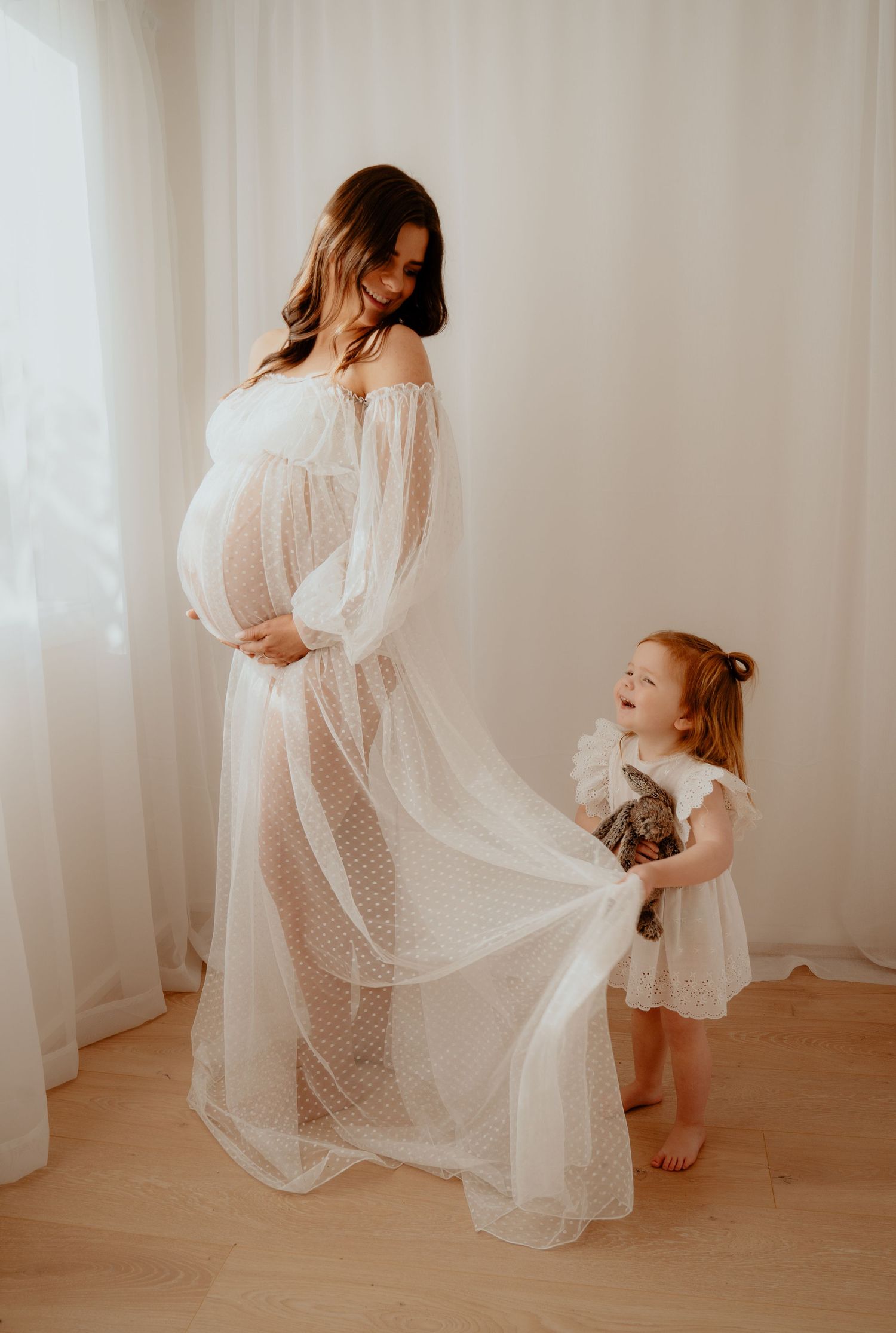 A pregnant woman in a sheer white maternity gown smiles as a little girl in a white dress holds her flowing skirt.