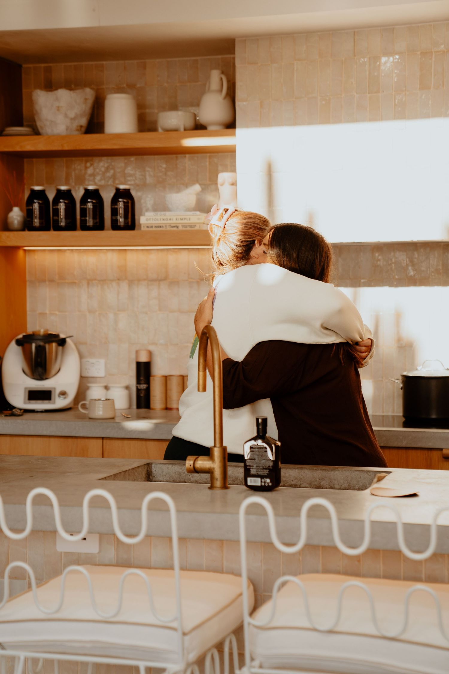 Two women embracing in a modern kitchen with warm beige tiles and open wooden shelves.