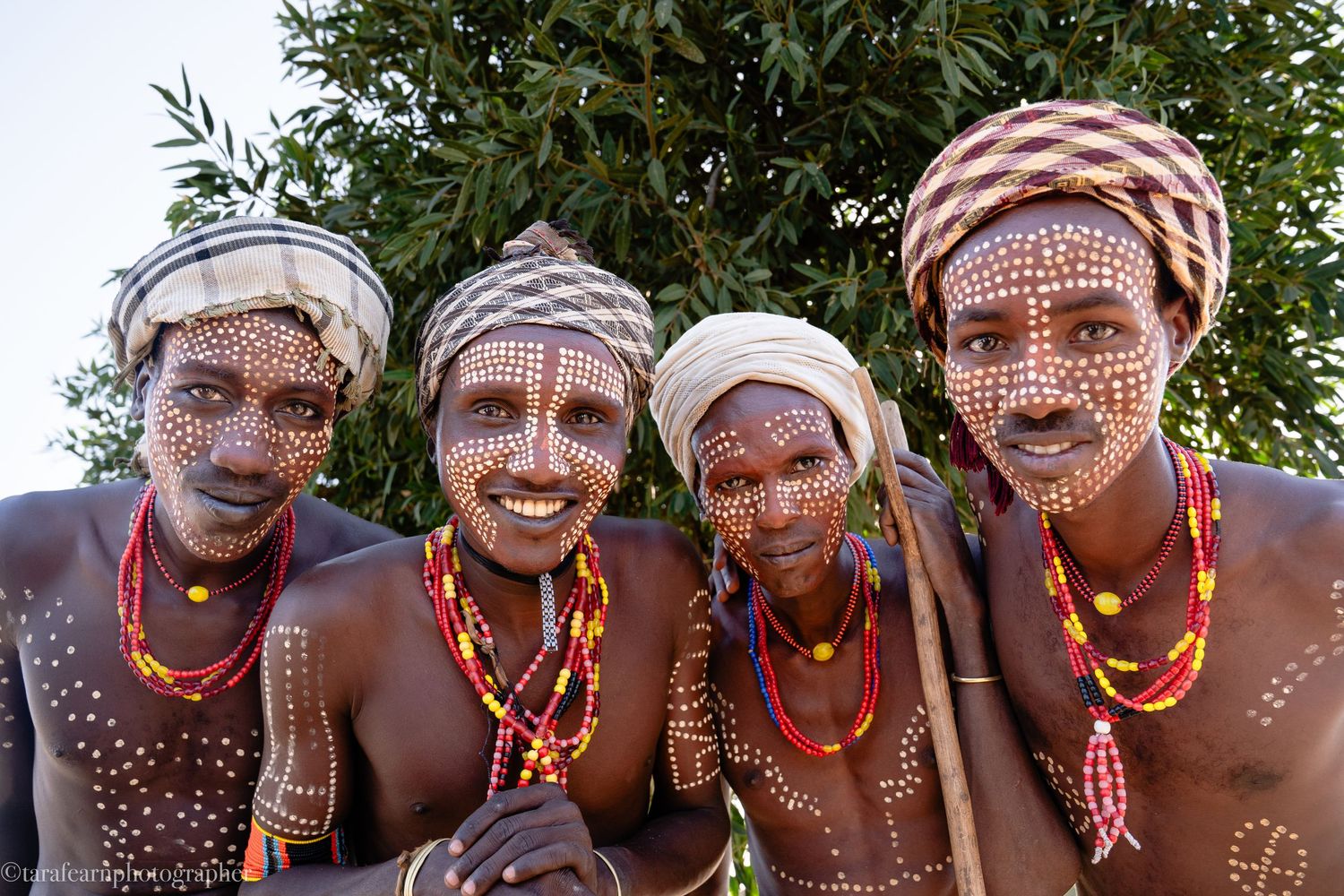 Four smiling African tribal men with white dot face paint and colorful beaded necklaces outdoors.