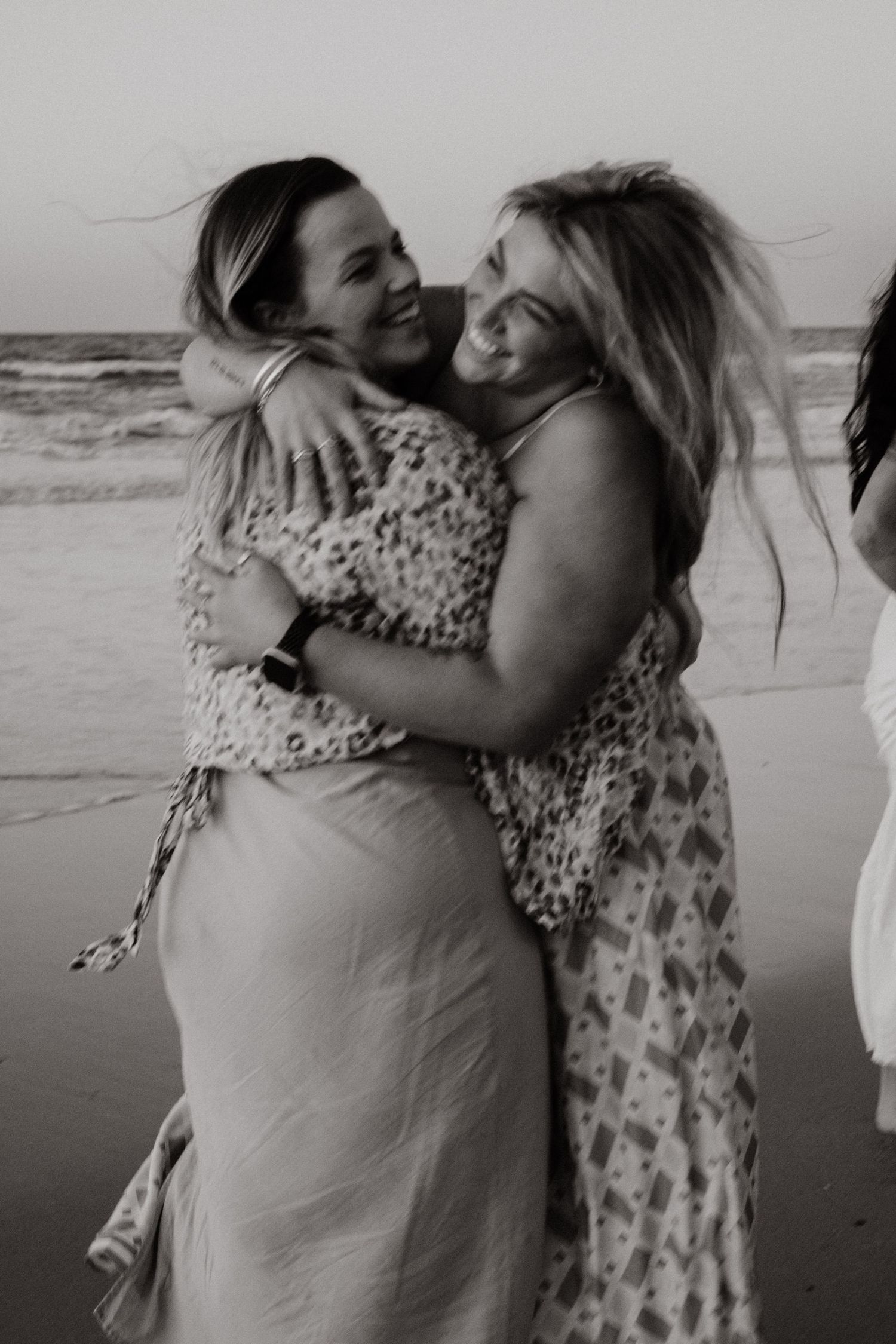 Black and white photo of two smiling women hugging on a beach at sunset.