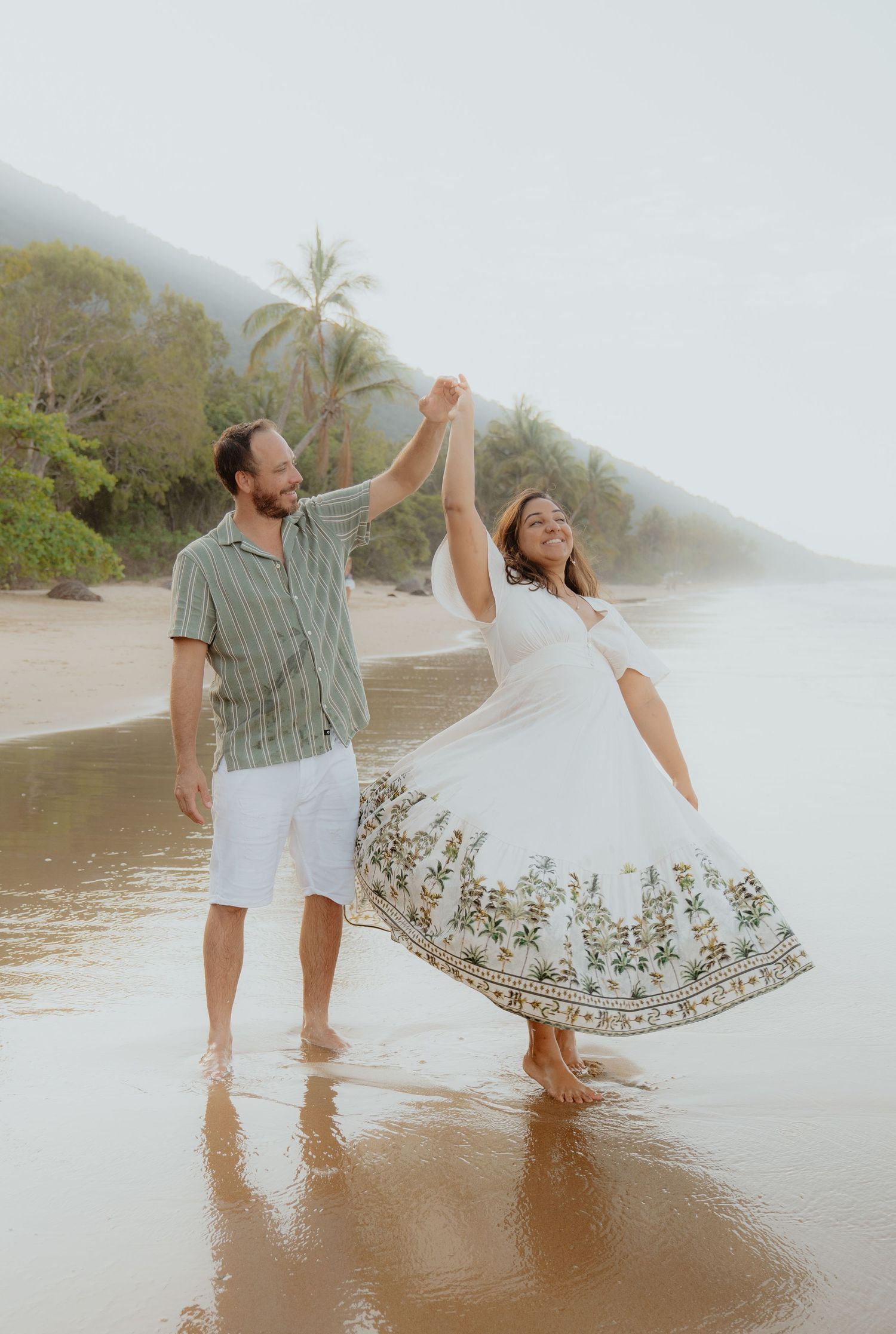 Couple dancing joyfully on a misty tropical beach, woman twirling in a floral white dress.