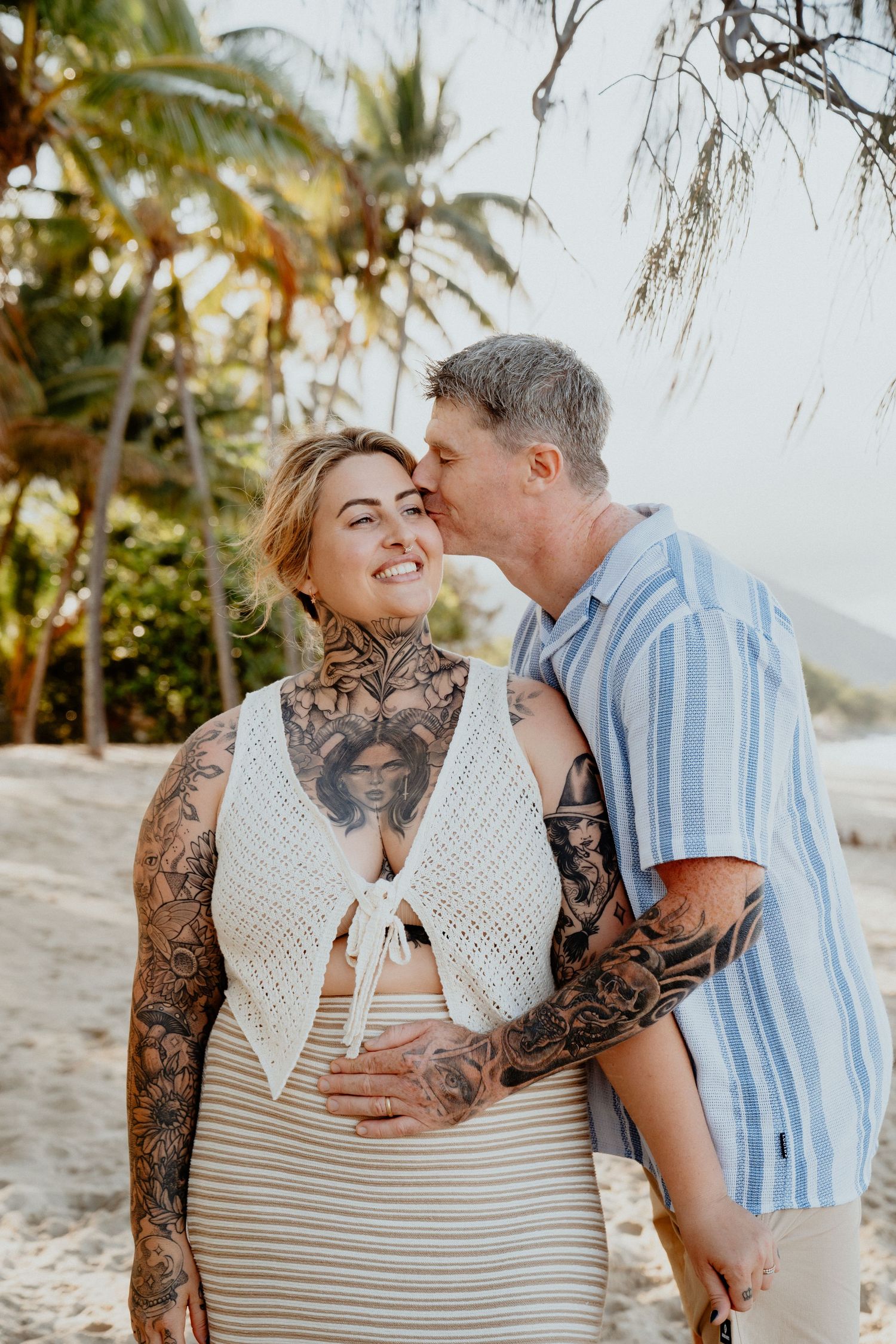 A tattooed couple shares a tender kiss on a tropical beach, with palm trees in the warm background.