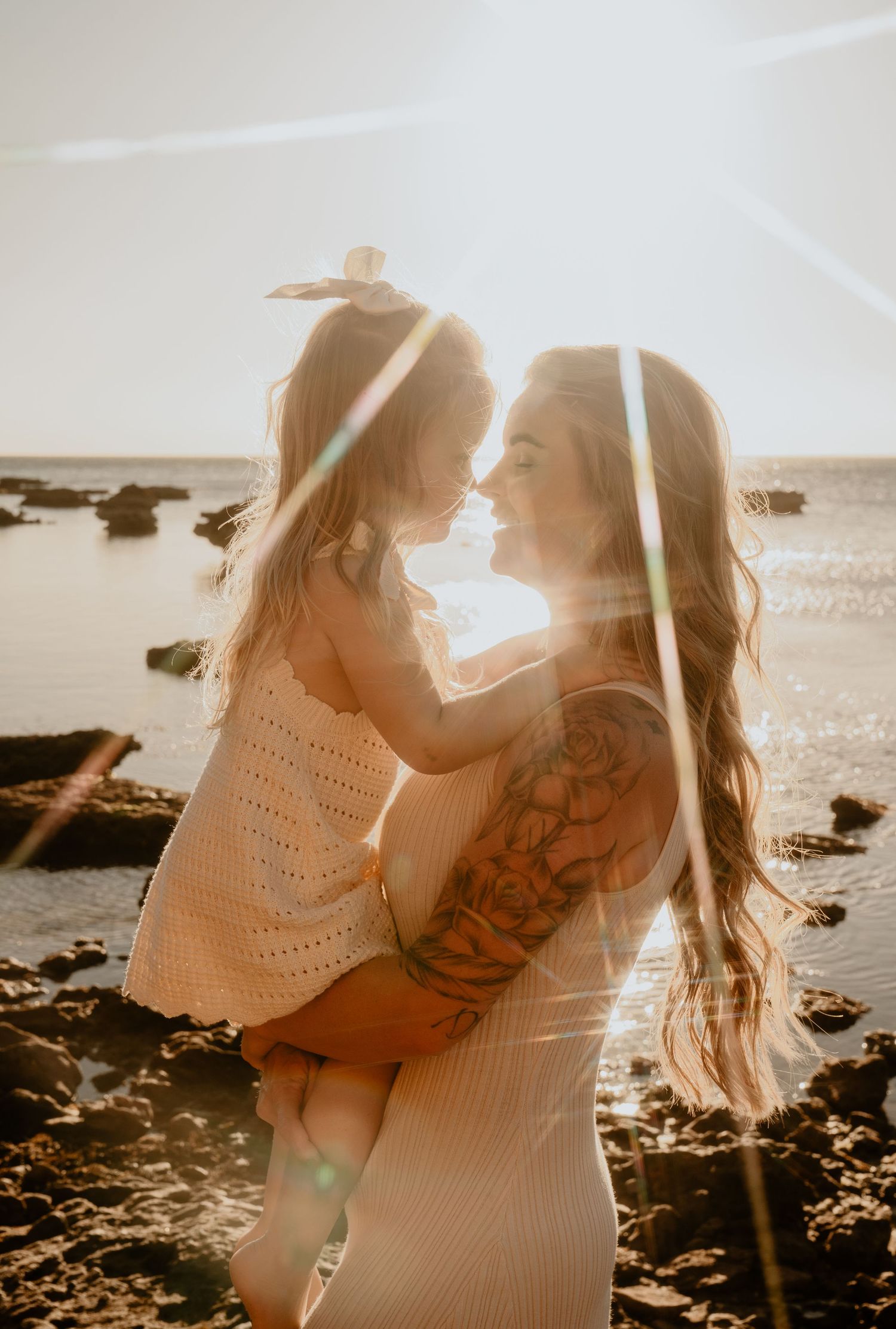 Mother with tattoos holding her young daughter face-to-face at sunset by the ocean.